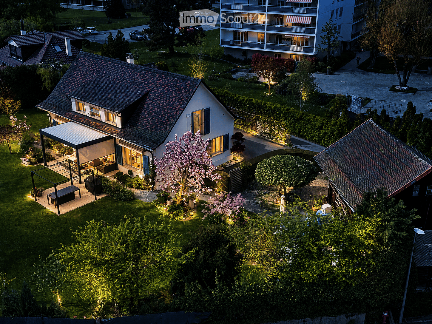 A modern two-story house with a tiled roof, surrounded by a well-maintained garden and illuminated pathways. The house features large windows and a covered outdoor seating area with a table and chairs. Nearby, there are additional residential buildings an