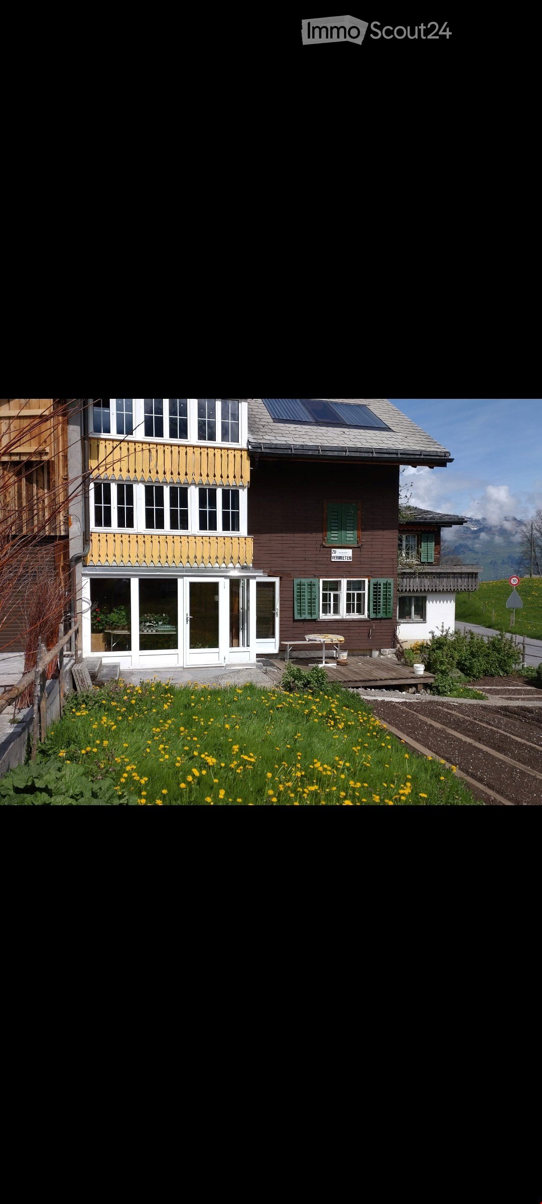 House. Two-story house with brown and yellow exterior, green shutters, solar panels on the roof, and a well-maintained garden with yellow flowers in the foreground.