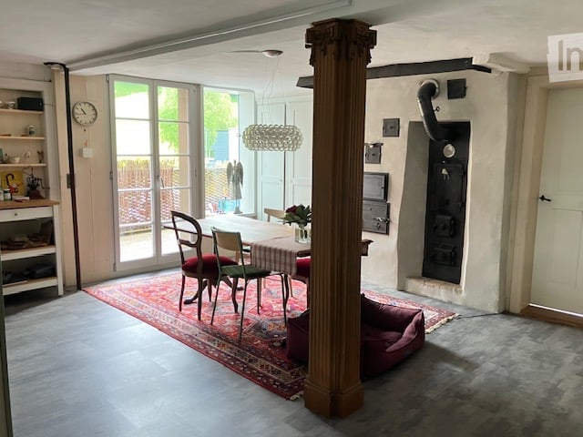 Furnished Room. Bright, open living space with a central dining table and chairs, a large area rug, a dog bed, a shelf with various items, and a clock on the wall. Large glass doors lead to an outdoor area.