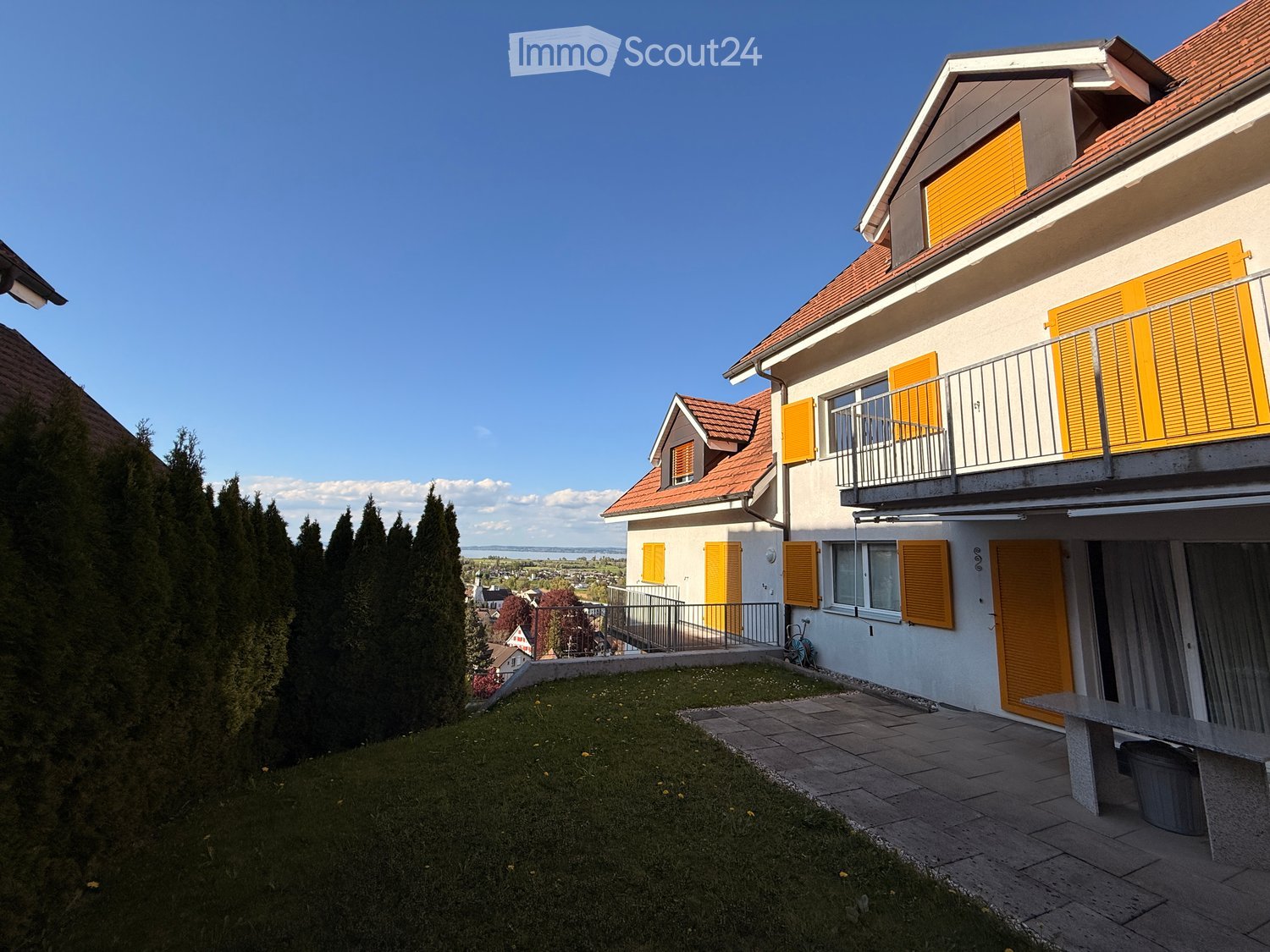 House. Two-story house with white walls, red-tiled roof, yellow shutters, and a balcony. The house is surrounded by a well-maintained lawn and trees, with a paved pathway leading to the entrance.