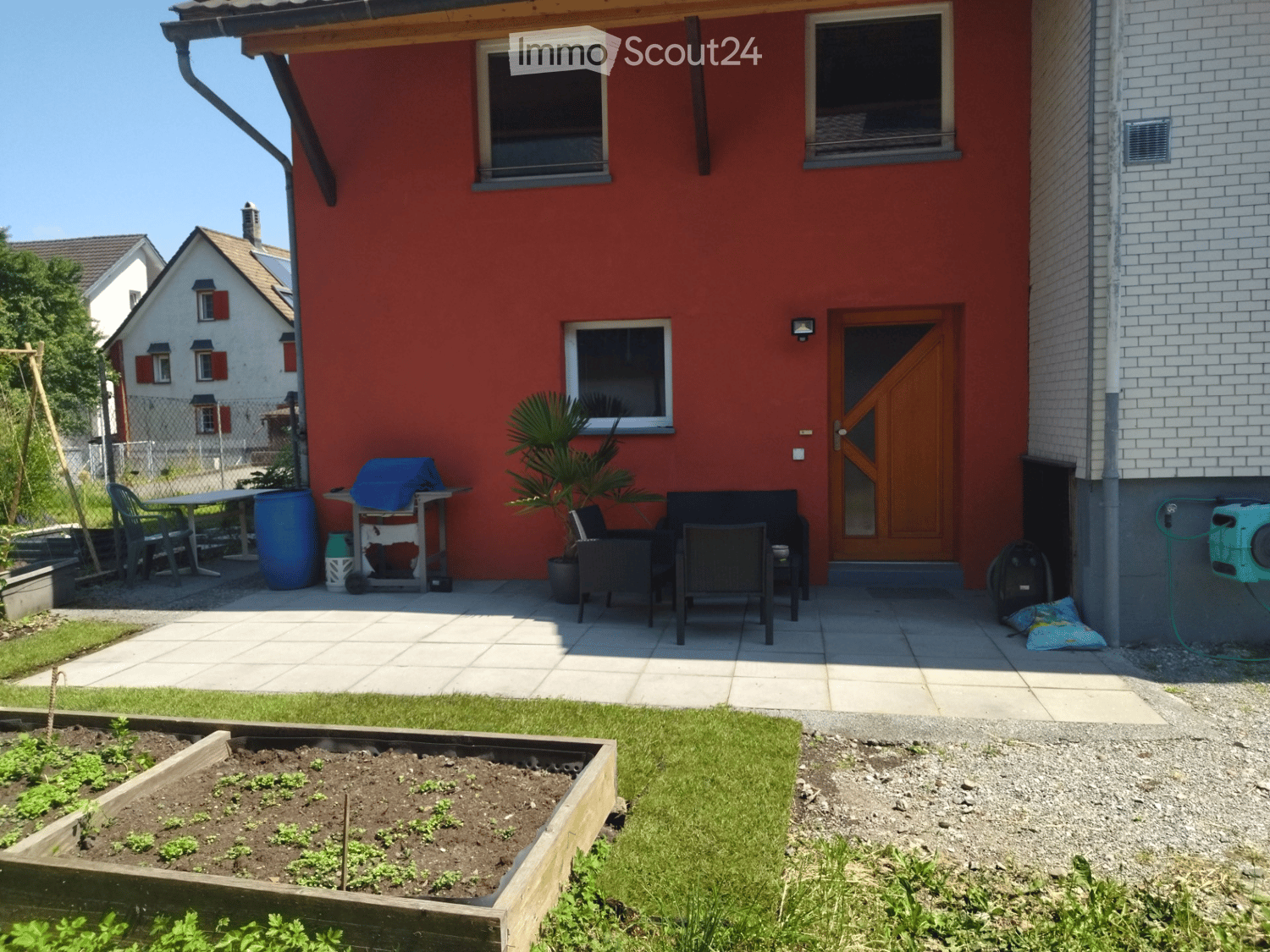 Garden area with raised wooden planter, patio seating, grill, and potted plant near the house entrance.