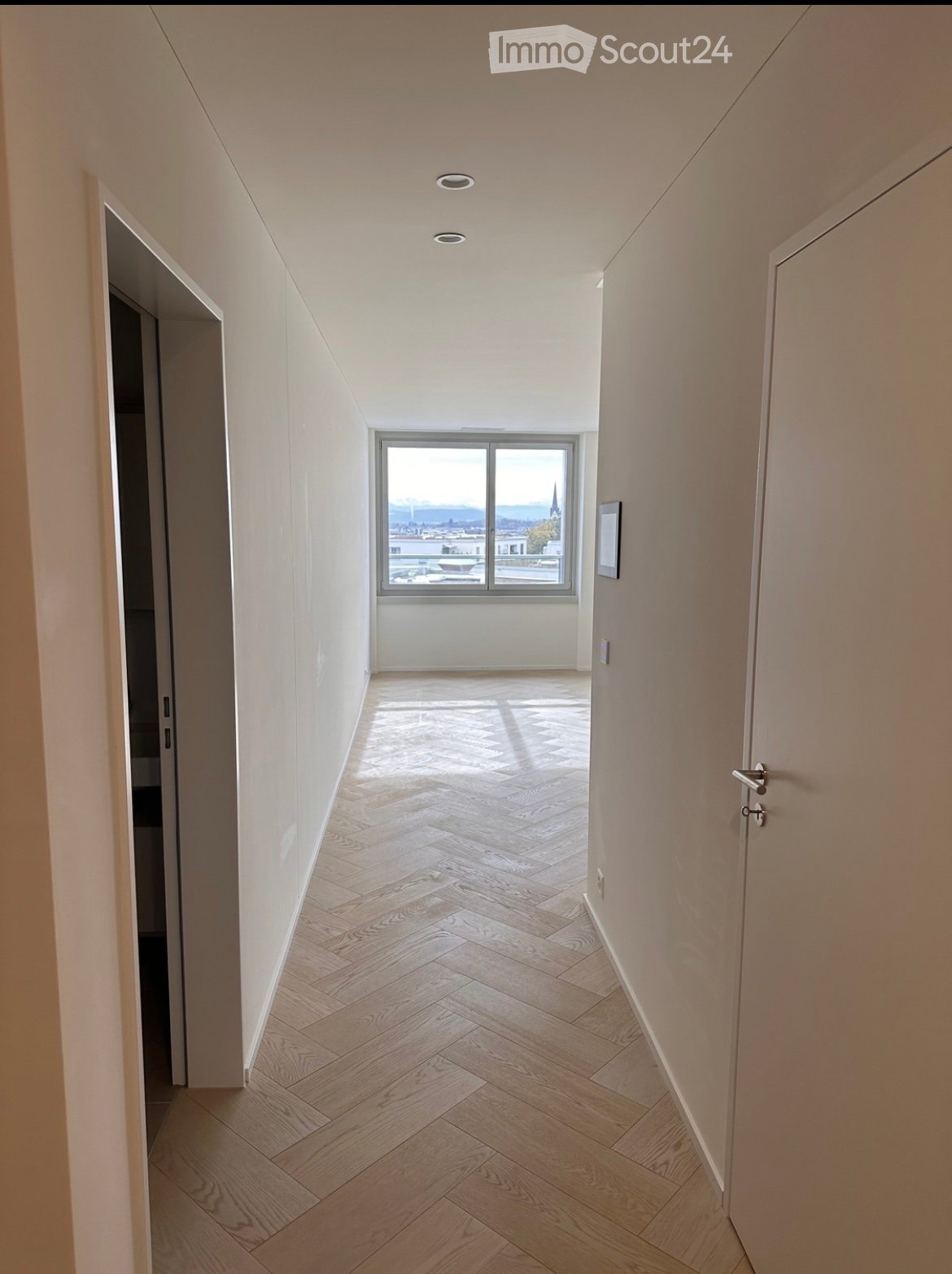 Corridor / Hallway. Modern hallway with herringbone wooden flooring, white walls, recessed ceiling lights, and a large window offering a city view.