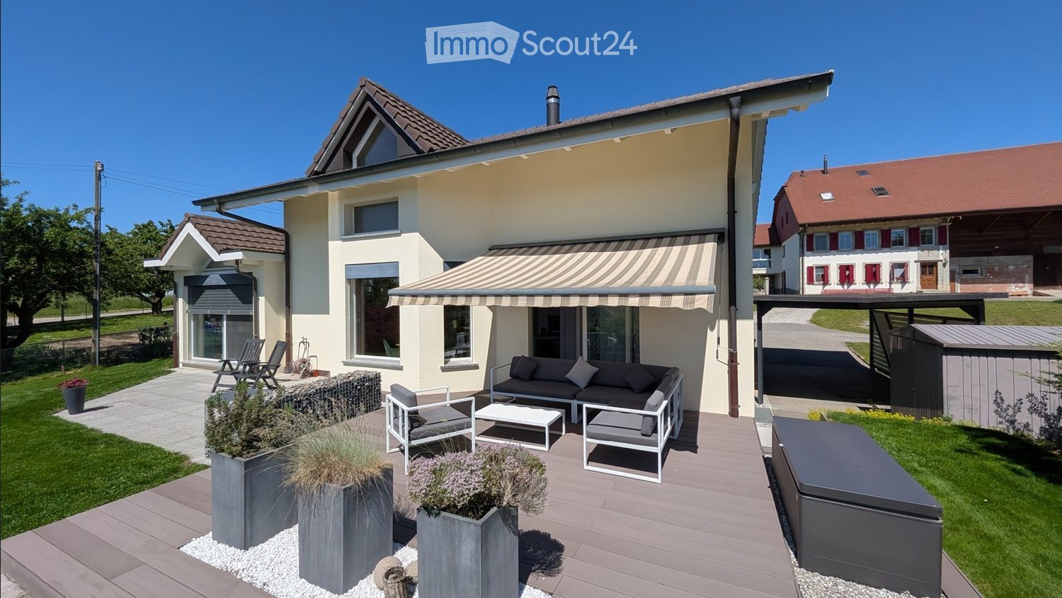 Two-story house with a modern design, featuring a covered patio area with outdoor seating, including a sofa, chairs, and a coffee table. The house has large windows, a striped awning, and is surrounded by a well-maintained garden with potted plants and a