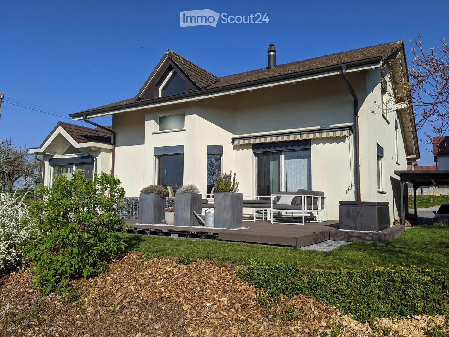 House. Modern two-story house with beige exterior, large windows, and a covered patio area with seating and planters.