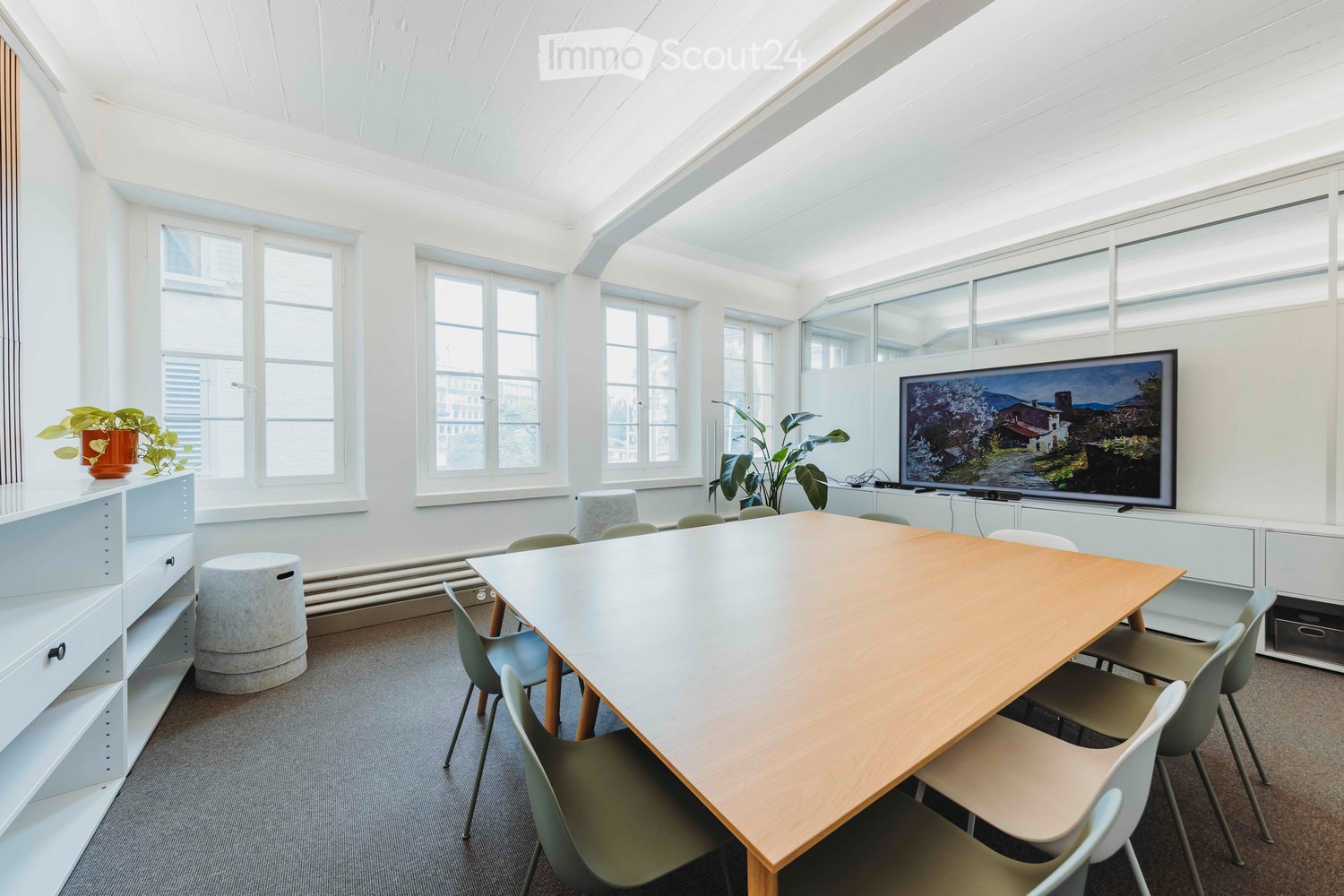 Furnished Room. Bright, modern meeting room with large windows, a long wooden table surrounded by chairs, a round stool, a large screen, and potted plants on shelves.