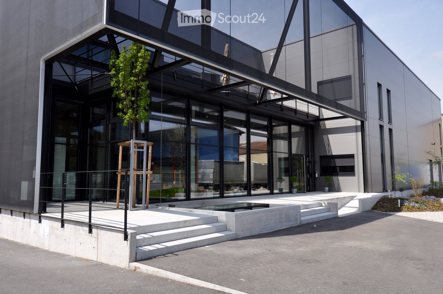 Building. Modern building with large glass windows and a sleek, contemporary design. Features a raised ground floor, small water feature, and small landscaped area with plants and trees.