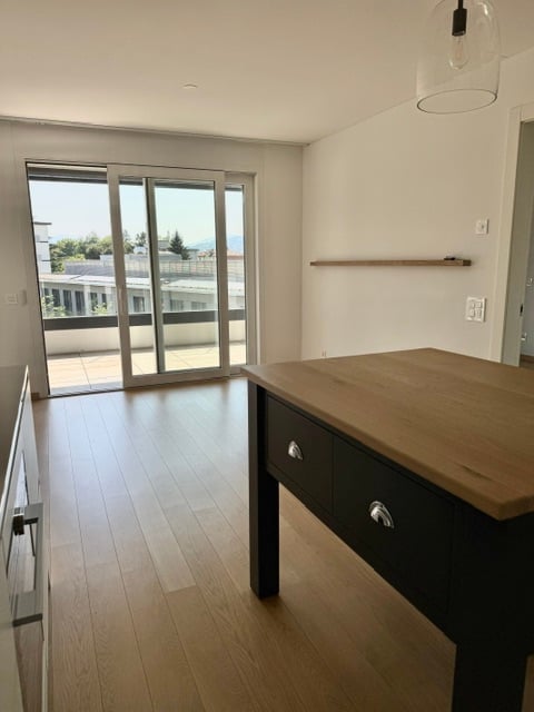 Bright, unfurnished room with wooden flooring, large sliding glass doors leading to a balcony, and a simple wooden table with drawers.