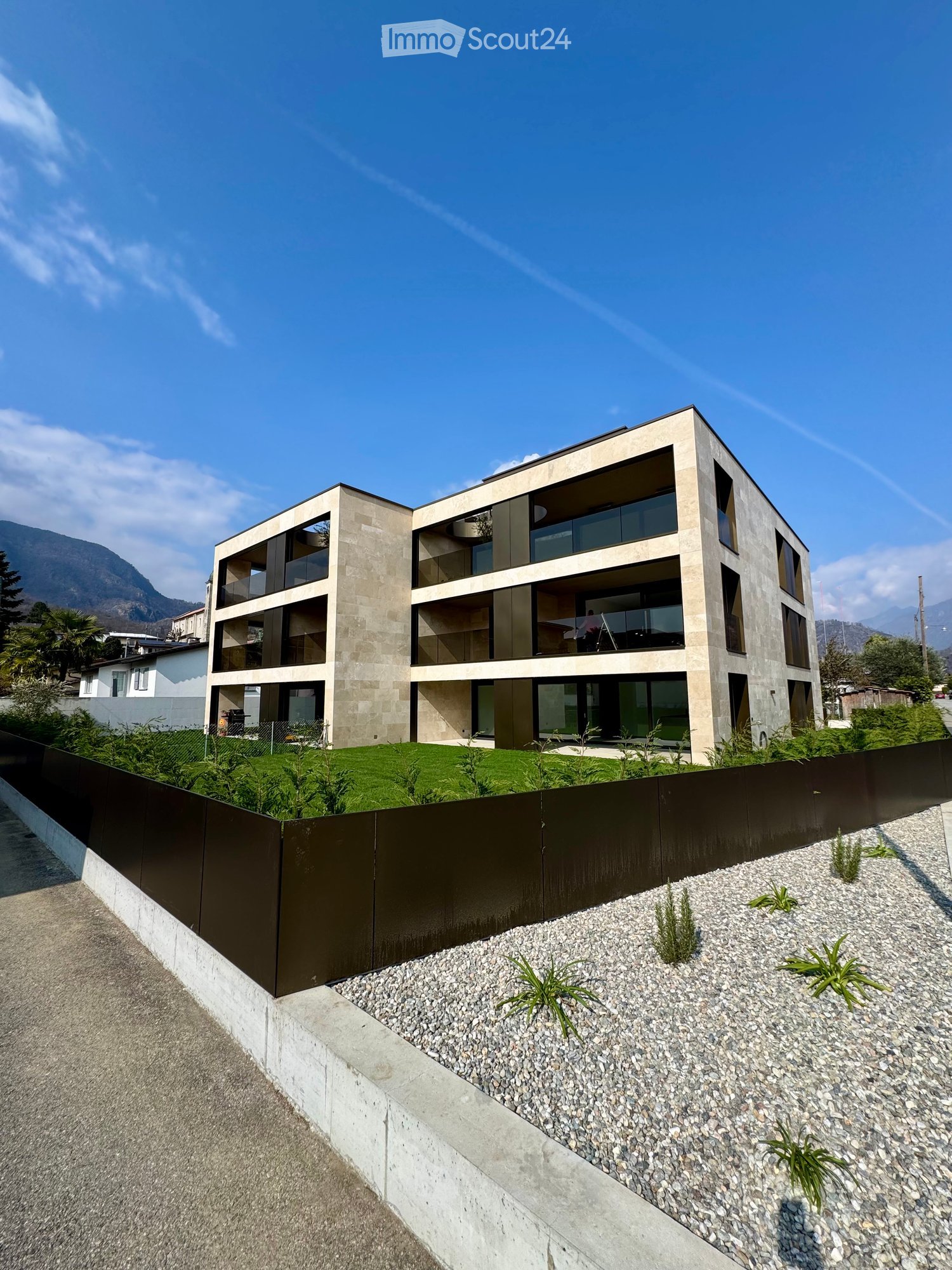 Building. Modern multi-story building with large windows and balconies, surrounded by a low fence and landscaped area with gravel and small plants.