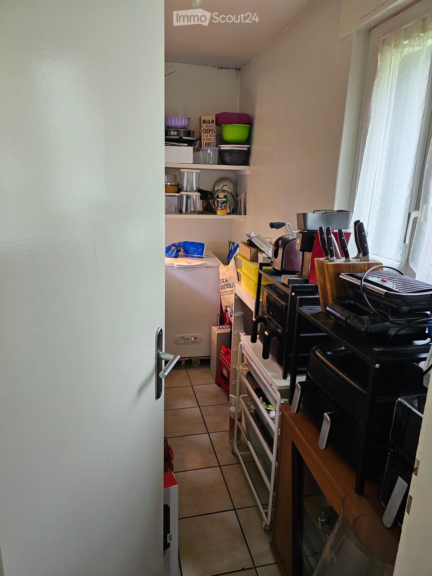 Kitchen. A small kitchen with tiled flooring, white walls, and various appliances including a refrigerator, microwave, toaster, and oven. Shelves hold bowls, plates, and cups. A wooden cutting board with knives is on the counter, and a small cart holds ad