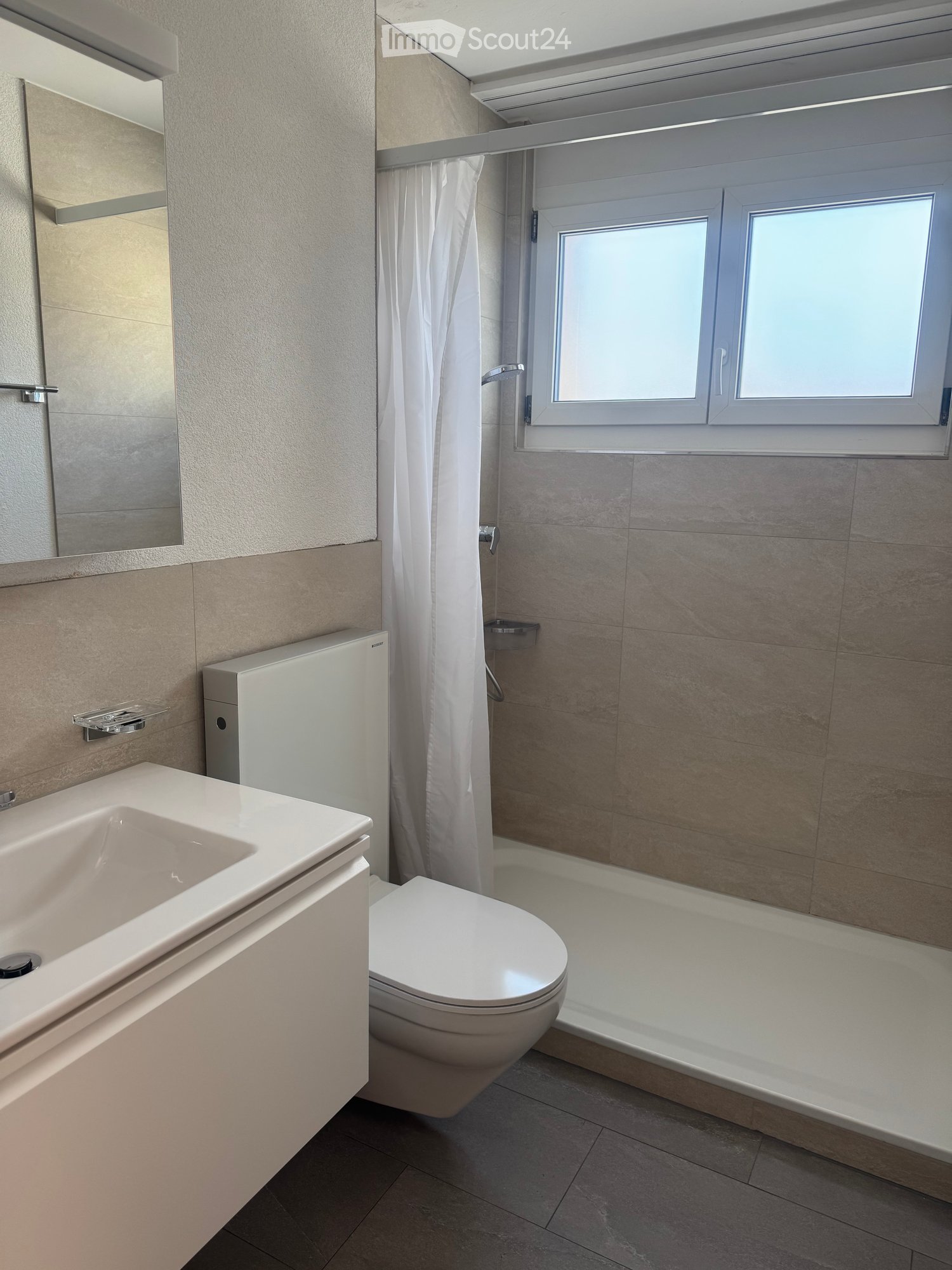 Bathroom. Modern bathroom with a white sink, toilet, mirror, and shower area with a curtain. Light-colored tiles and a window provide natural light.