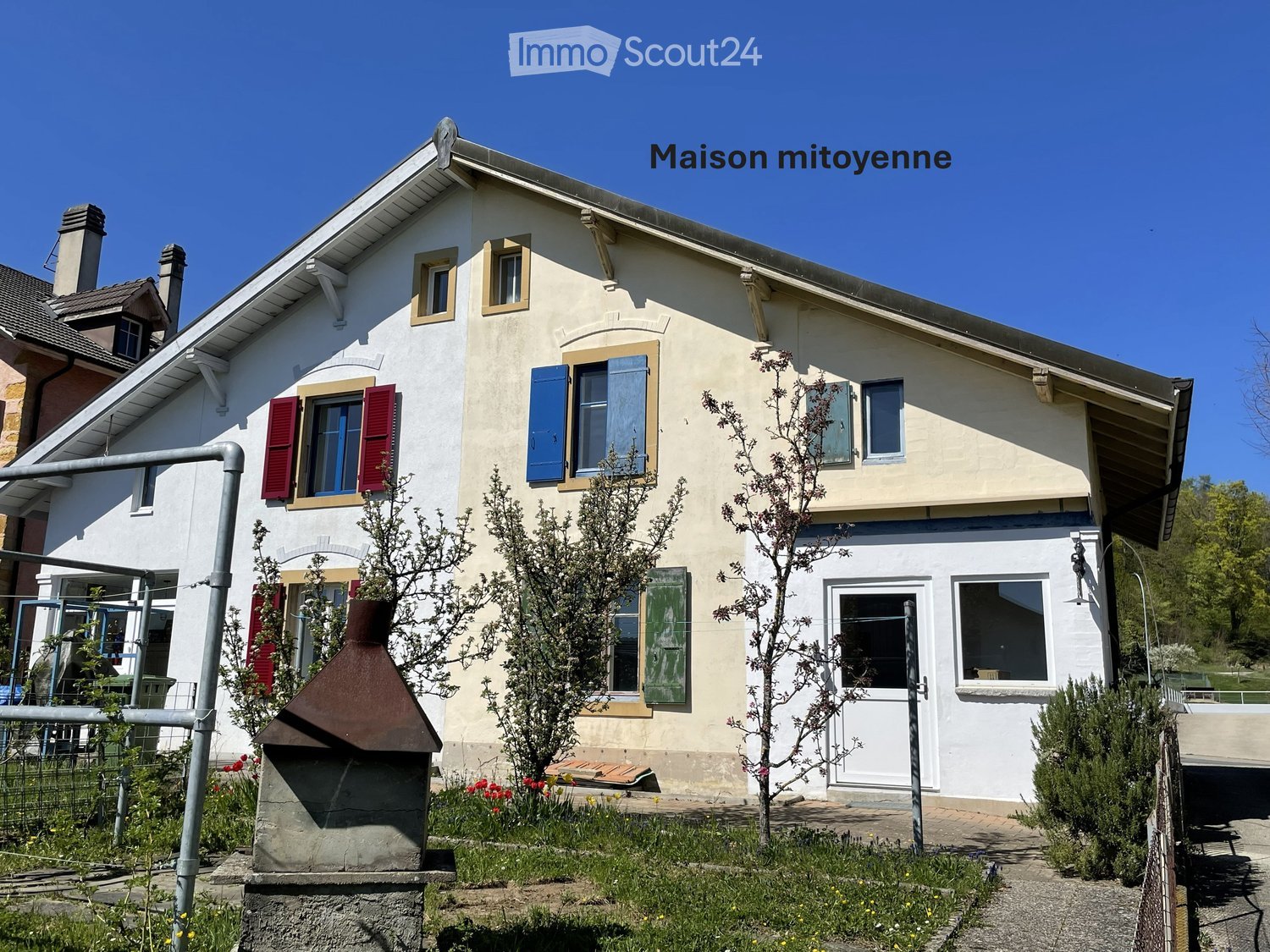 House. Two-story house with beige walls, red shutters, and blue shutters, surrounded by small trees and shrubs.