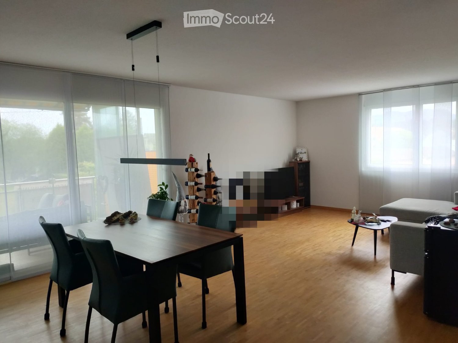 Furnished Room. Open-plan living and dining area with wooden flooring, large windows with blinds, a dining table with chairs, a sofa, a coffee table, a wine rack, and a television.