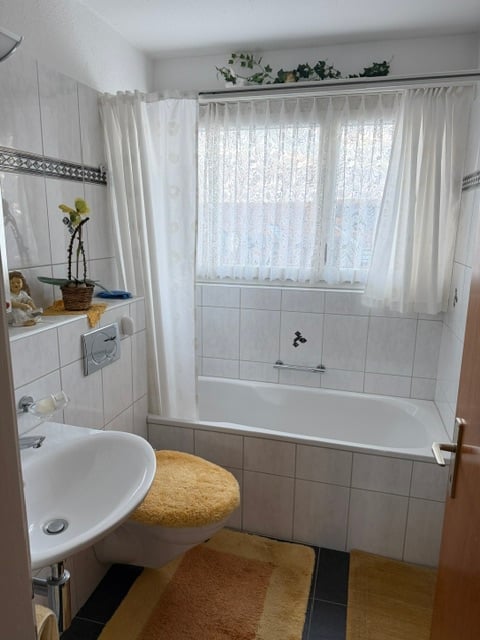 Bathroom. Bathroom with a white bathtub, toilet with a yellow seat cover, sink, mirror, and a window with a white curtain. A small plant is placed on the windowsill.