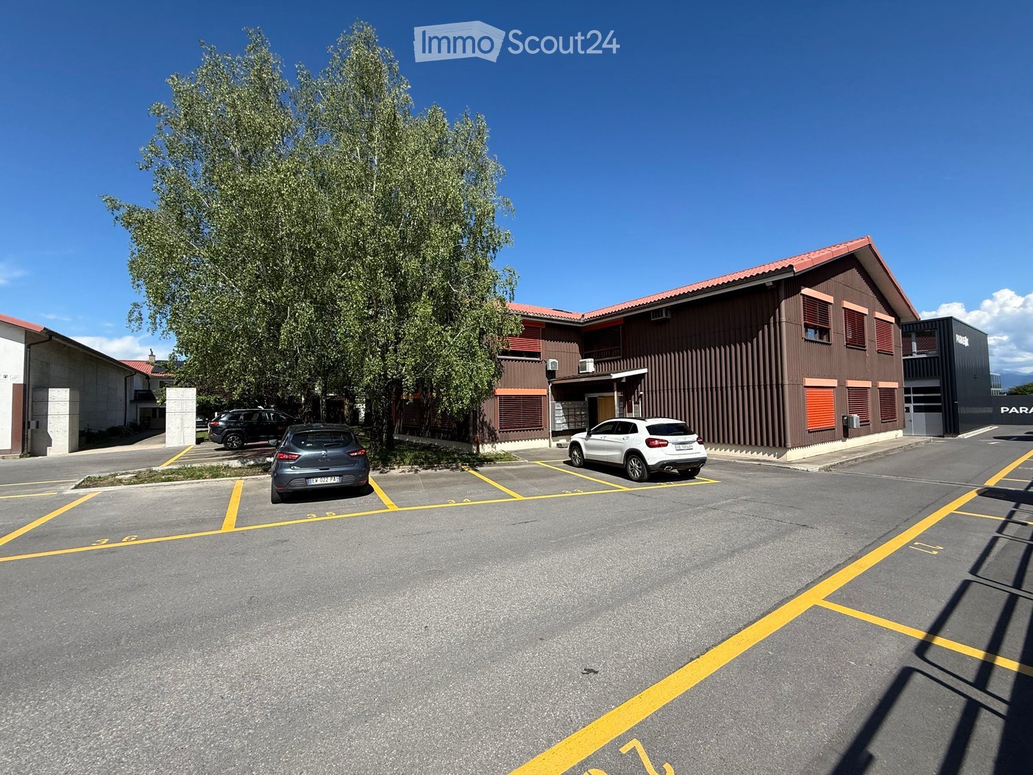 Building. A modern two-story building with a brown facade and red-tiled roof, surrounded by a parking lot with several parked cars and a large tree.