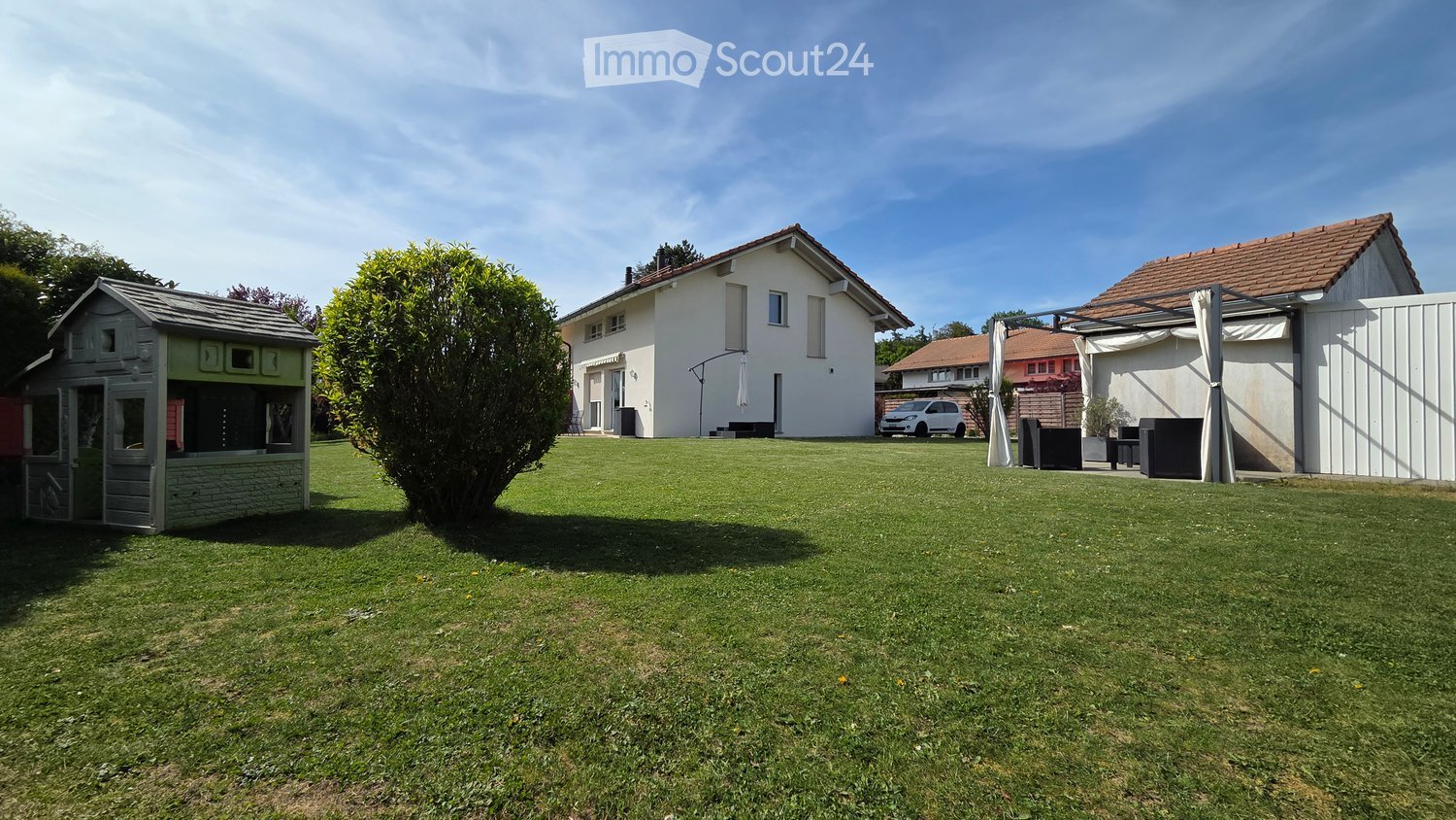 Garden / Outdoor Area. Well-maintained lawn with a playhouse, a large tree, and a white car parked near a house with a covered patio area.