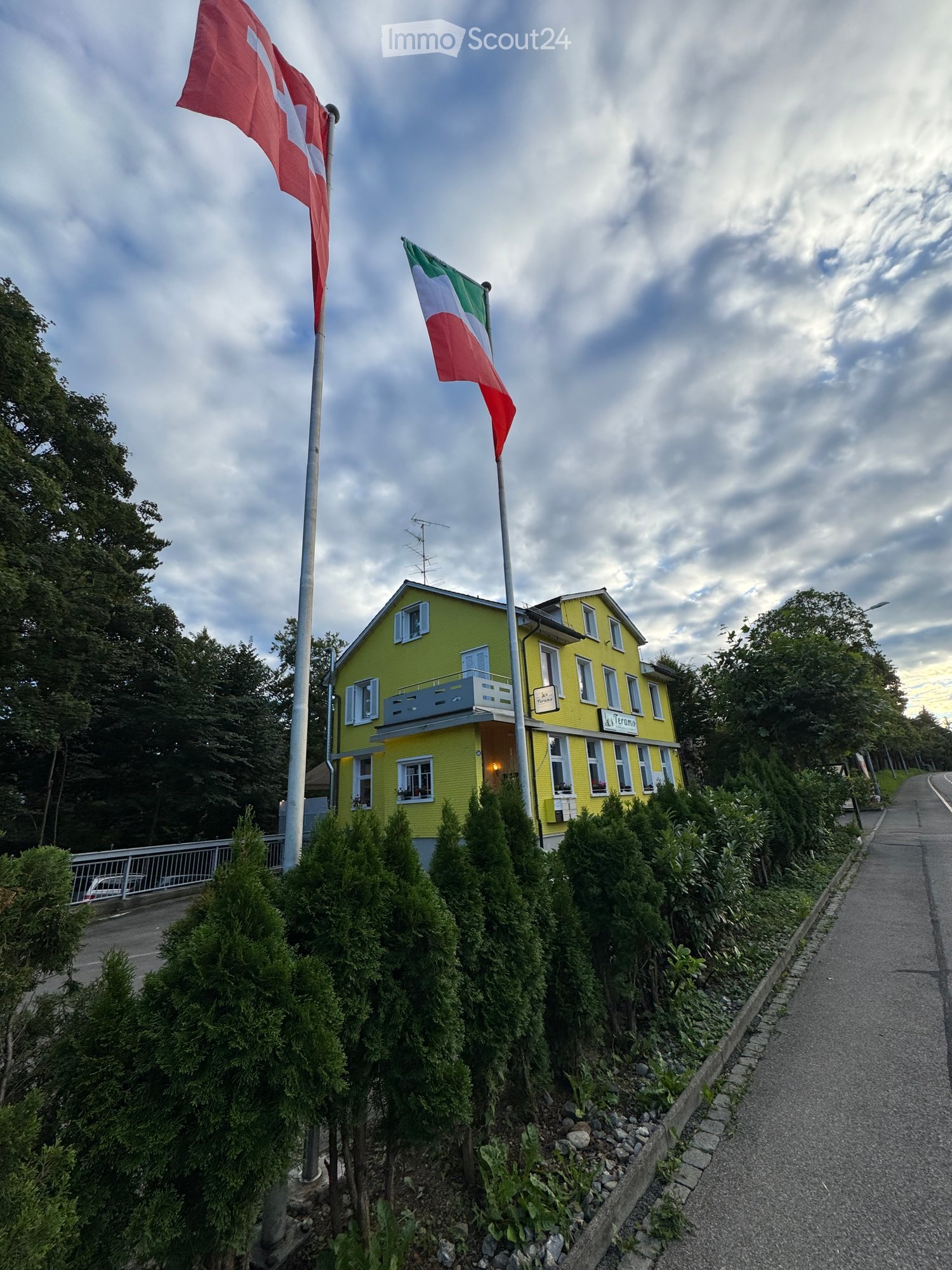 Building. Bright yellow two-story building with white trim, multiple windows, and a small balcony. Two flags (Swiss and Italian) are displayed on poles in front of the building. The building is surrounded by neatly trimmed green bushes and trees, with a p