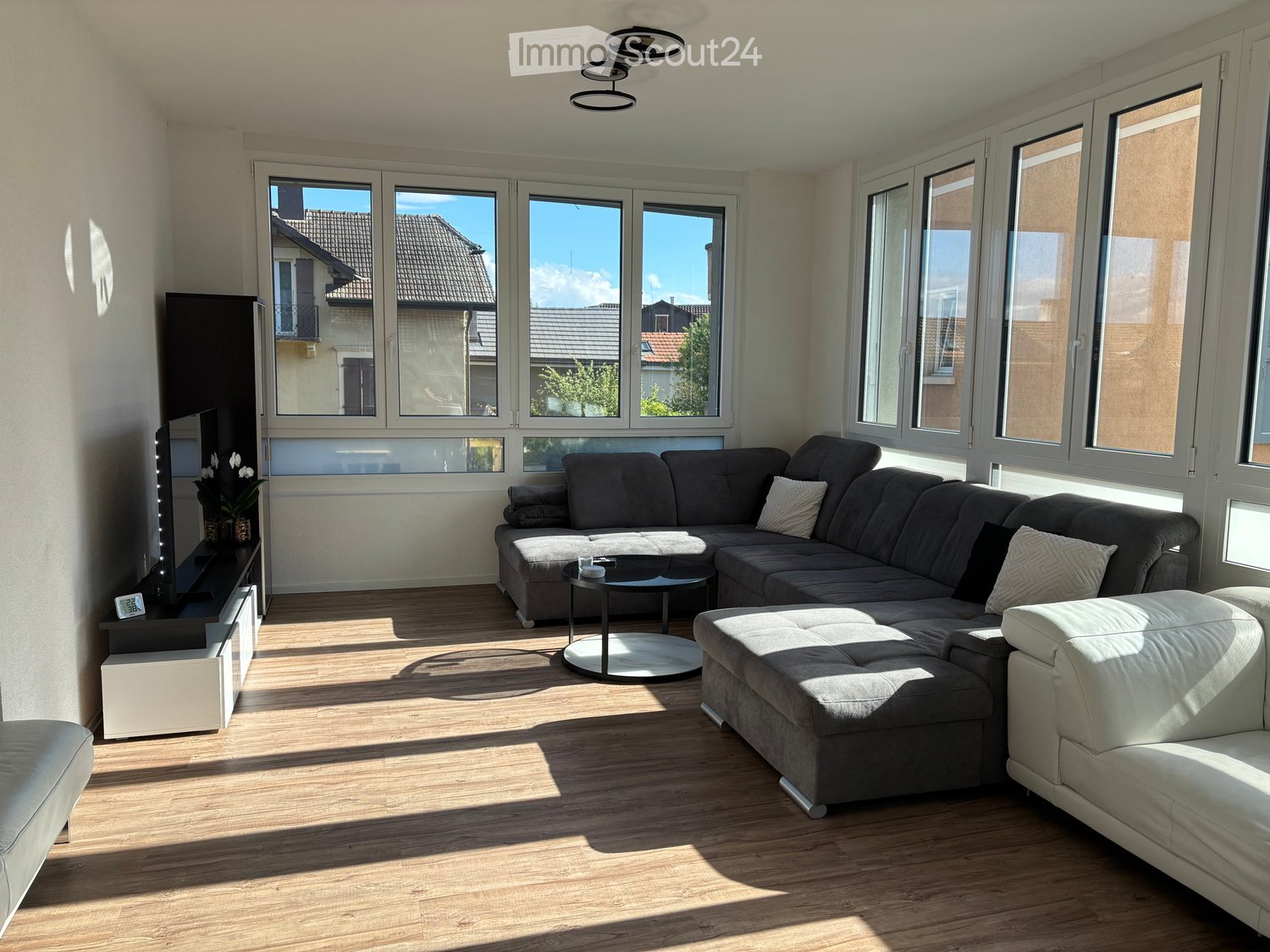 Living Room. Bright living room with large windows, grey sectional sofa, coffee table, ottoman, TV stand, and flower vase.