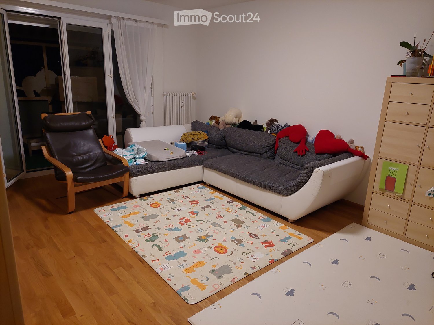 Living Room. Living room with a grey sectional sofa, leather armchair, wooden floor, colorful children's rugs, and a wooden drawer unit.
