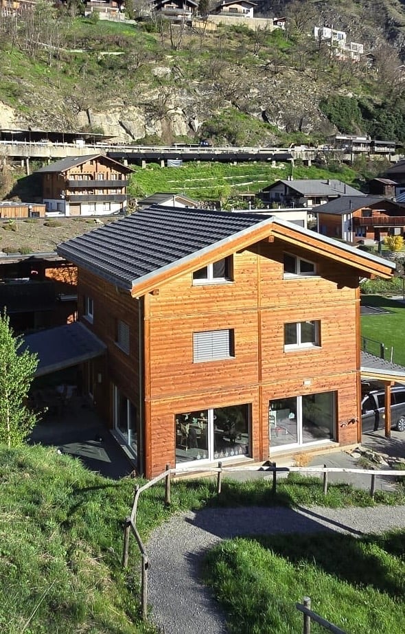 House. A modern wooden house with a dark grey roof, large windows, and a covered entrance area. The house is situated on a grassy slope with a gravel pathway leading to it. The surrounding area includes other houses and a scenic mountainous backdrop.