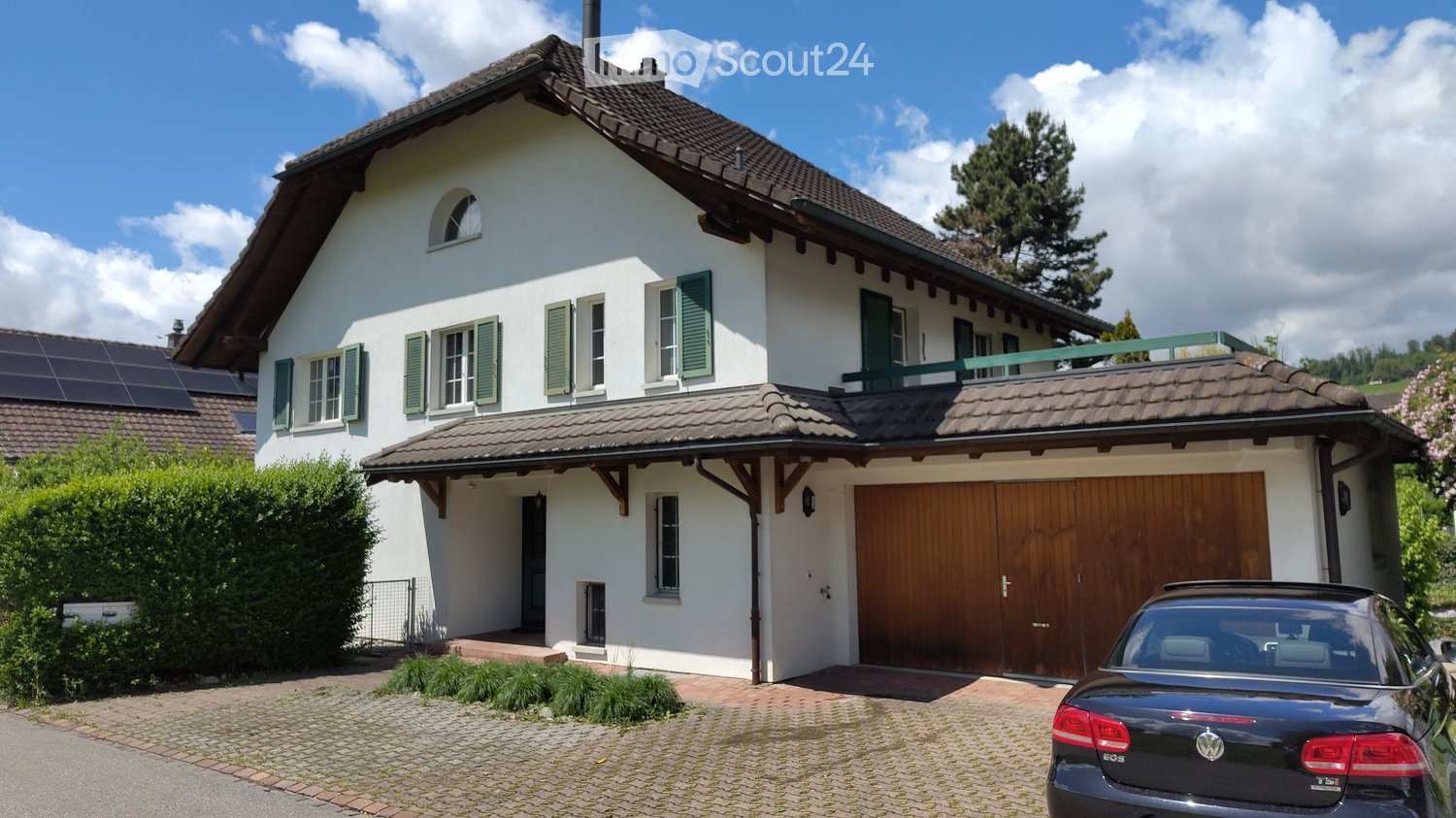 House. Two-story white house with green shutters, wooden garage, and solar panels on the roof. The house features a balcony on the upper level and is surrounded by greenery.