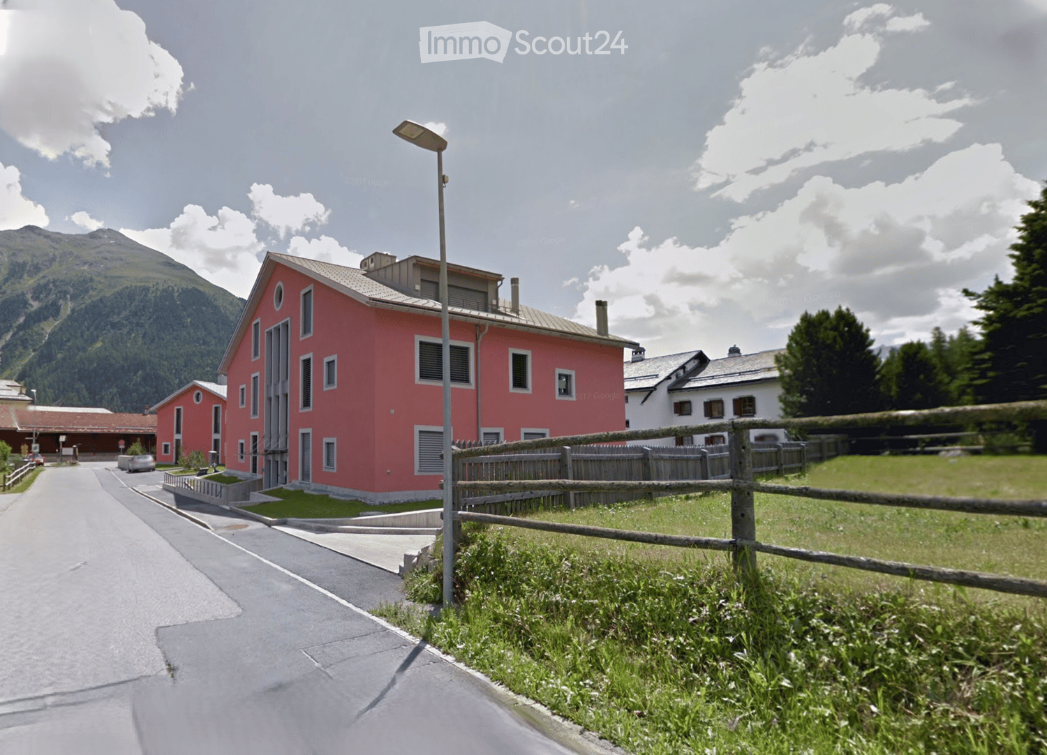 Building. A two-story red building with white-framed windows and a gray roof, situated in a rural area with a wooden fence and green grass in the foreground, with mountains visible in the background.
