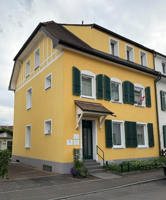 House. Two-story yellow house with white trim, green shutters, and a small porch with a flag. The entrance has a staircase leading to a green door.