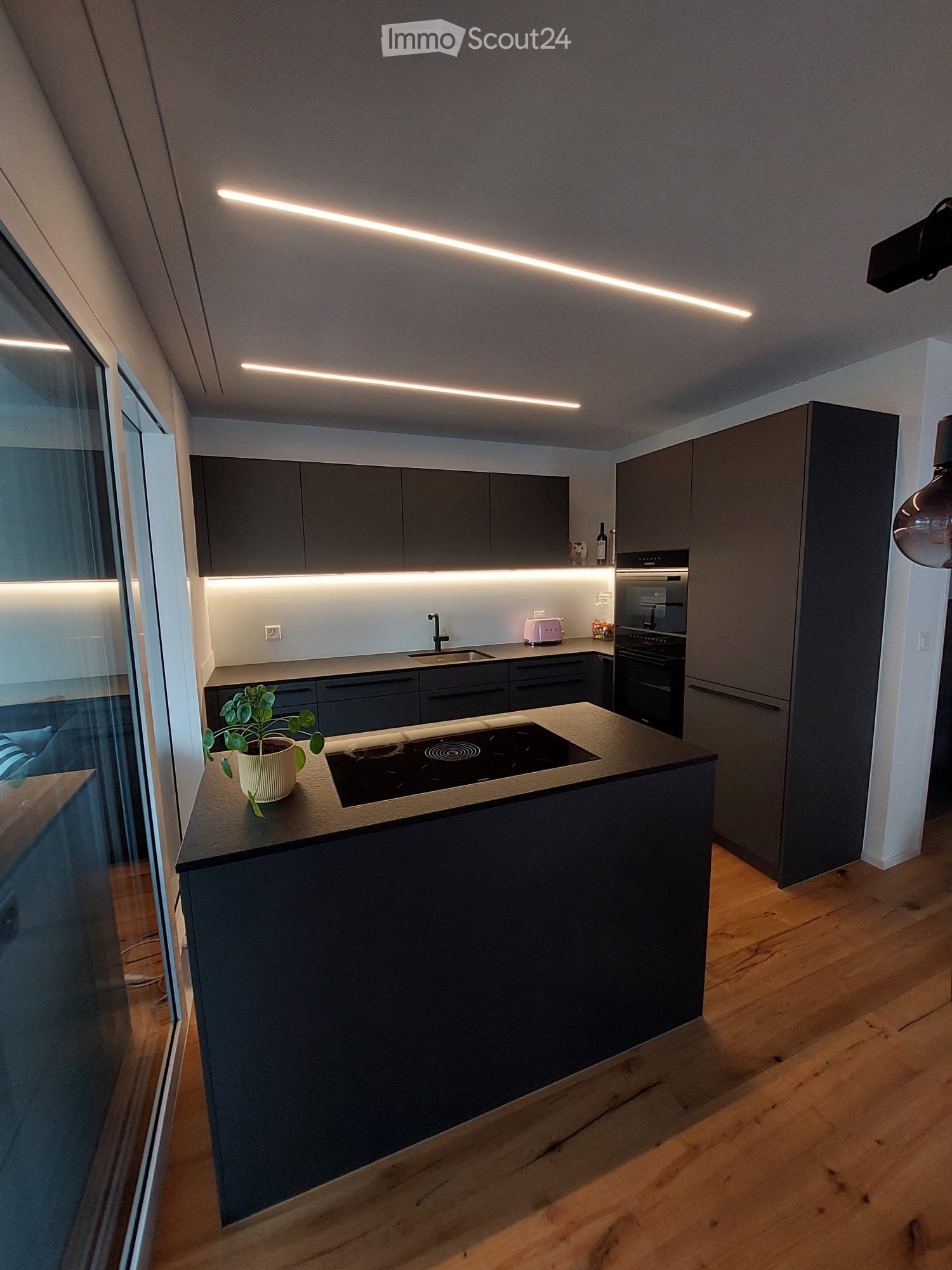 Kitchen. Modern kitchen with dark grey cabinets, a central island with an induction cooktop, and built-in appliances including an oven and a sink. The space features wooden flooring and sleek, contemporary lighting.