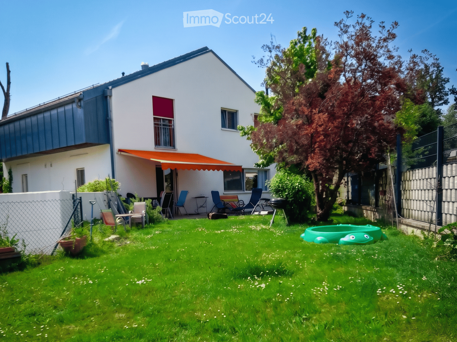 Garden / Outdoor Area. A well-maintained garden with a green lawn, patio furniture, a small inflatable pool, and a modern house with an orange awning in the background.