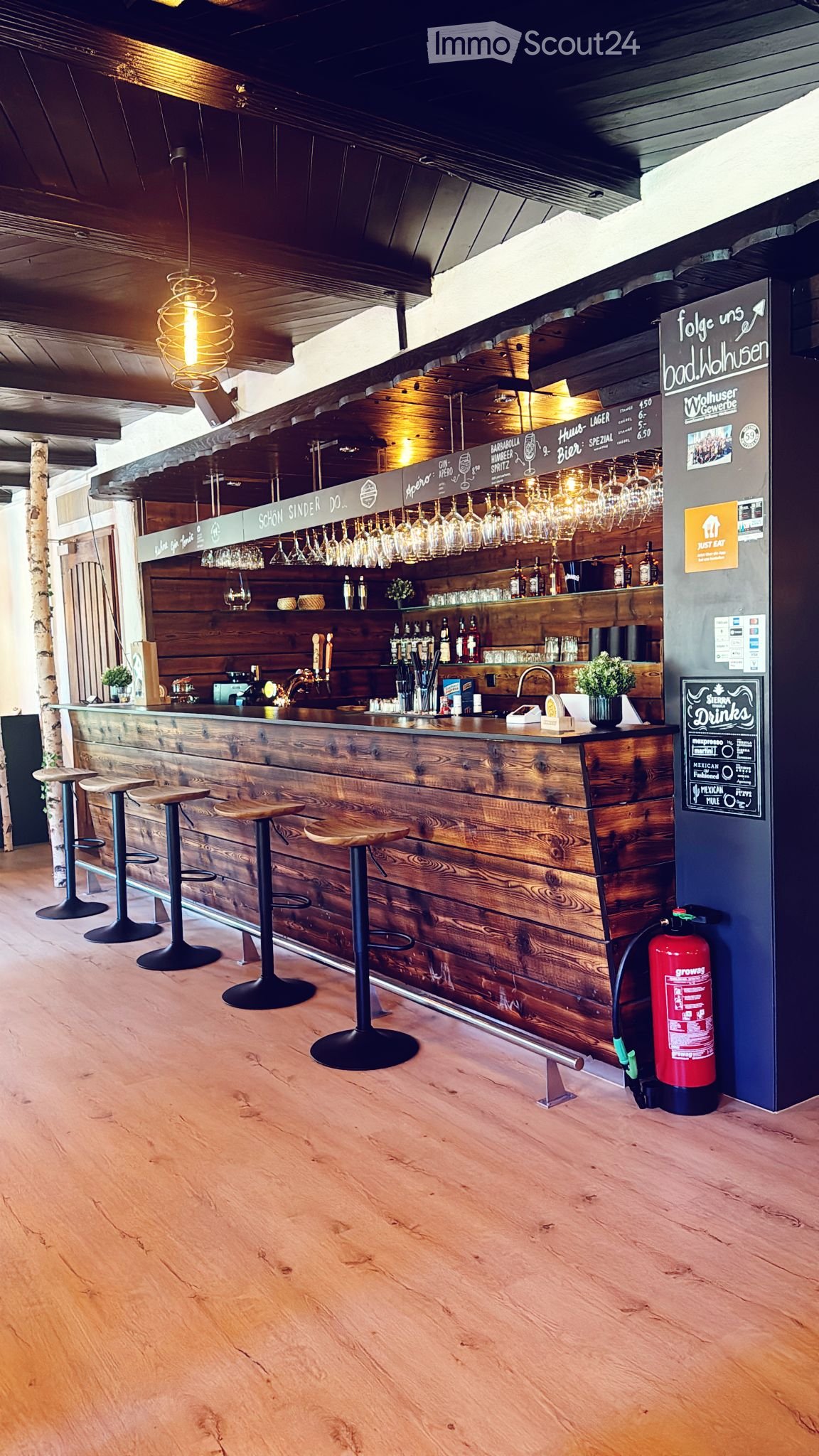 Wooden bar counter with hanging wine glasses, bottles, and stools, illuminated by hanging lamps.