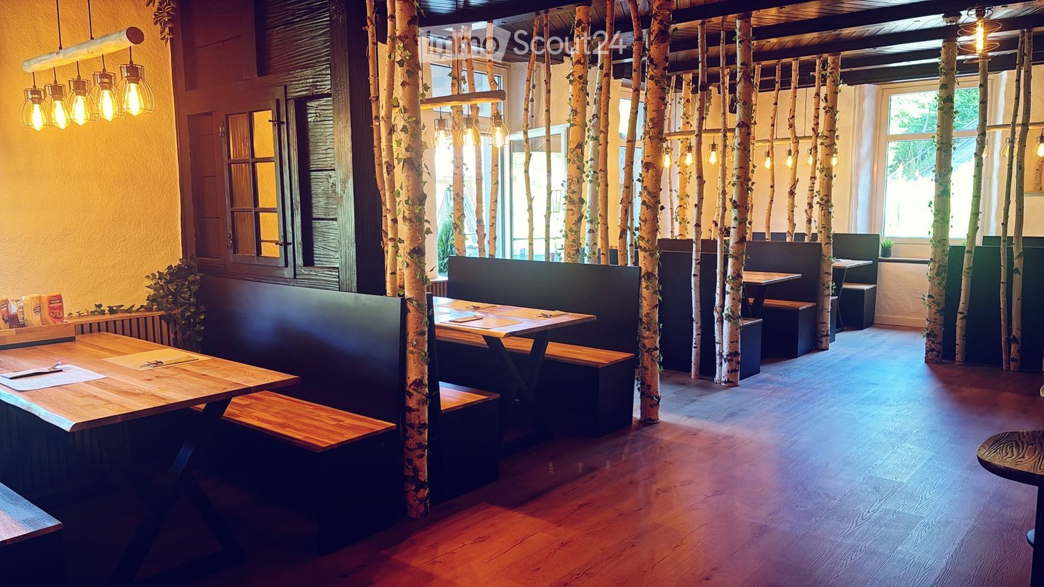 Restaurant interior with wooden tables, benches, and hanging lamps, featuring birch tree trunks as decorative columns.