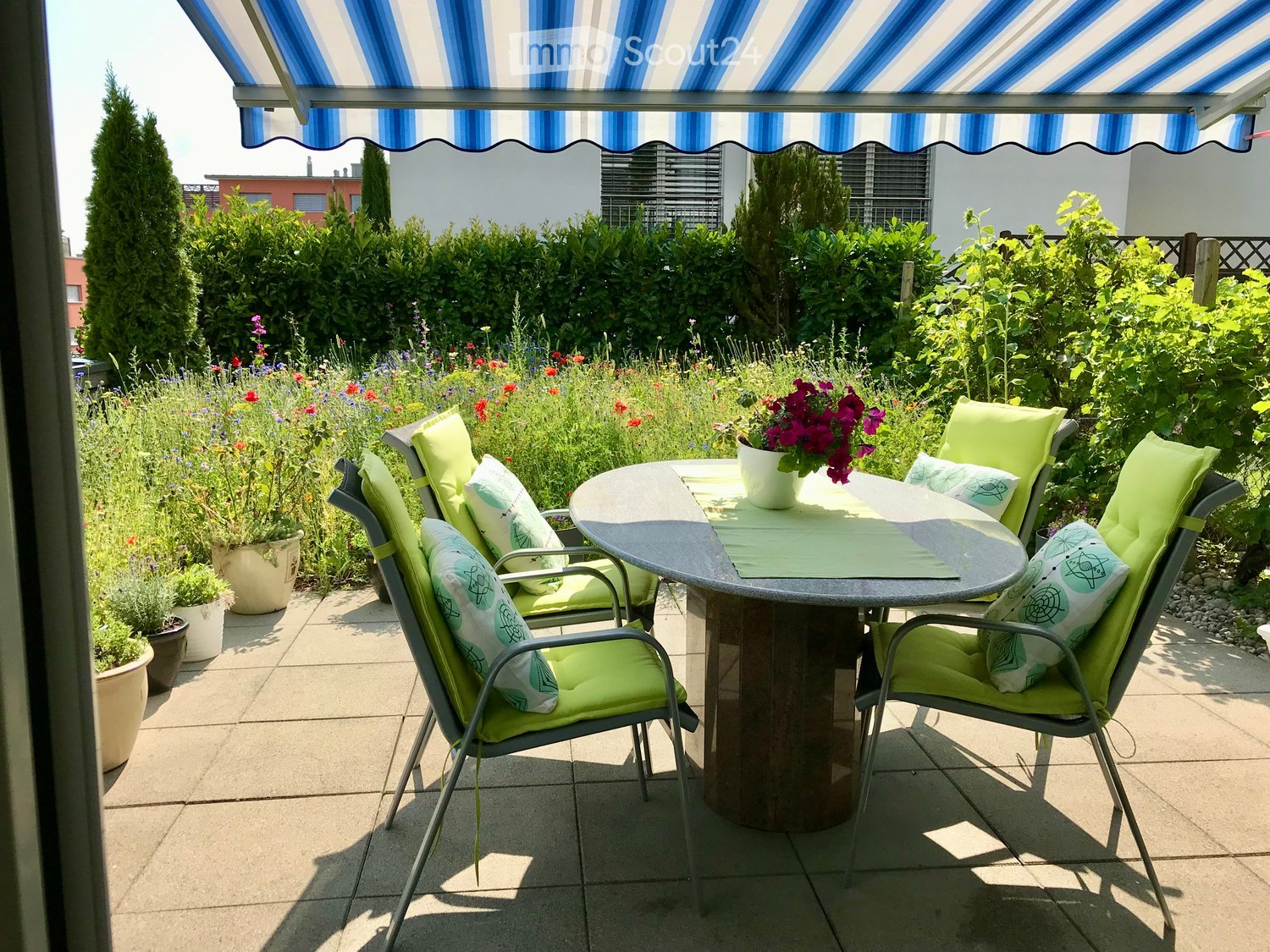 Garden / Outdoor Area. A sunny outdoor patio with a round table, four green chairs, and a flower pot on the table, surrounded by a garden with colorful flowers and greenery.
