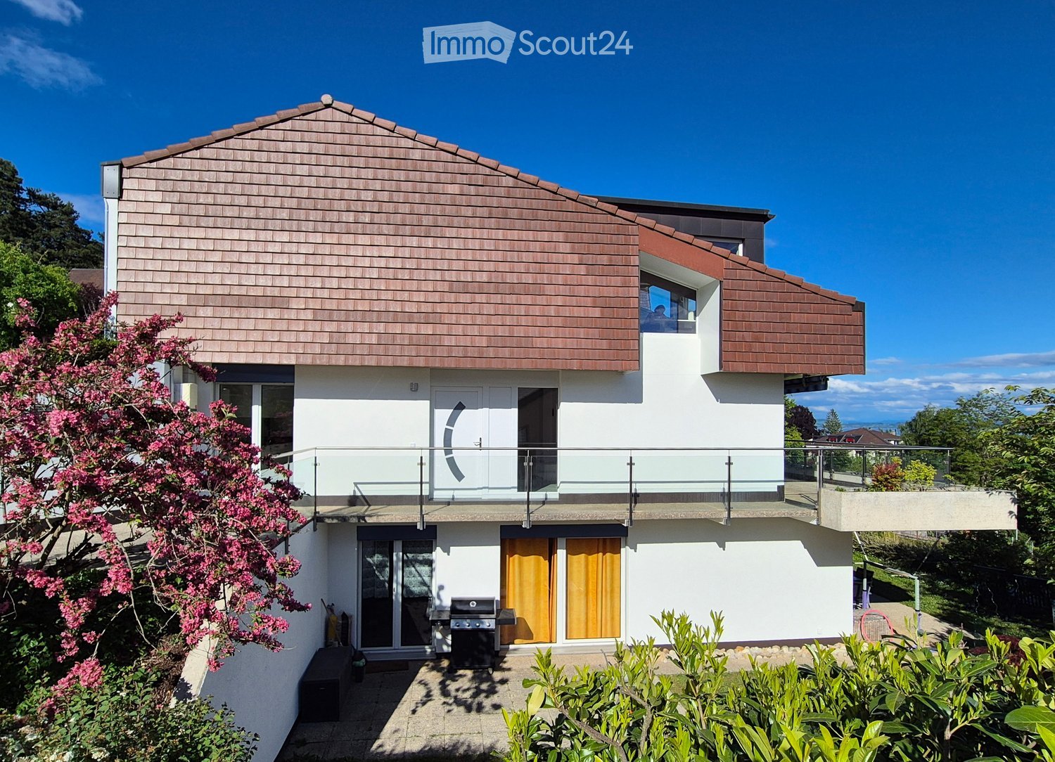 House. Modern two-story house with a sloped roof, white walls, and large windows. Features include a balcony, a raised ground floor, and a view of the ocean in the background.