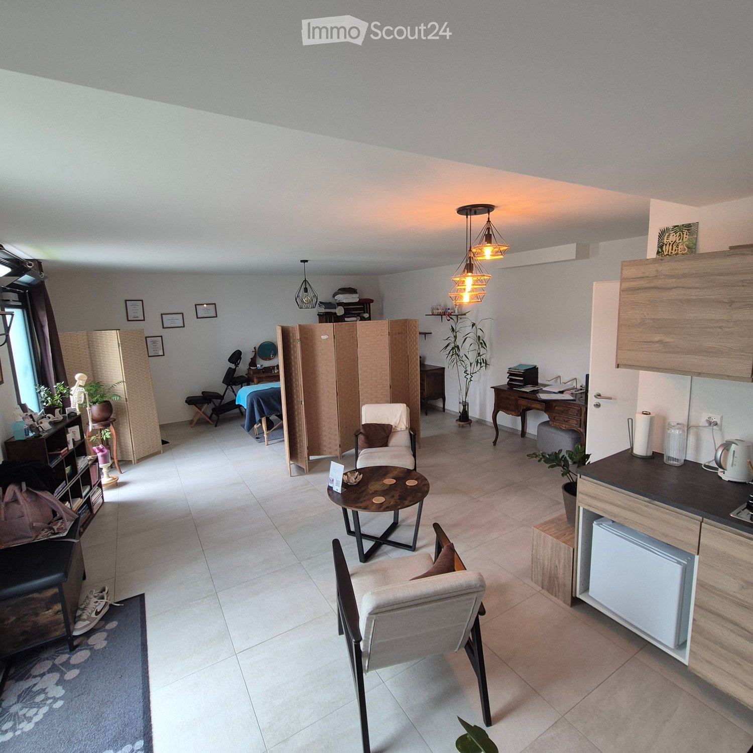 Furnished Room. The image shows a well-lit, modern living room with a mix of furniture and decor. There is a white sofa with a brown pillow, a small round wooden table with a few items on it, and several chairs arranged around the space. A kitchen area is