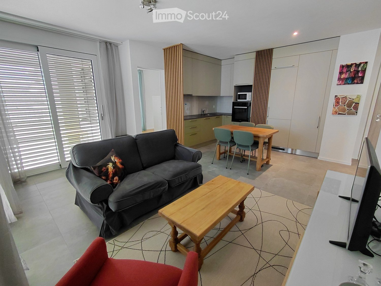 Furnished Room. Open-plan living and dining area with a grey sofa, wooden coffee table, dining table with chairs, and a small kitchen area with cabinets and appliances.