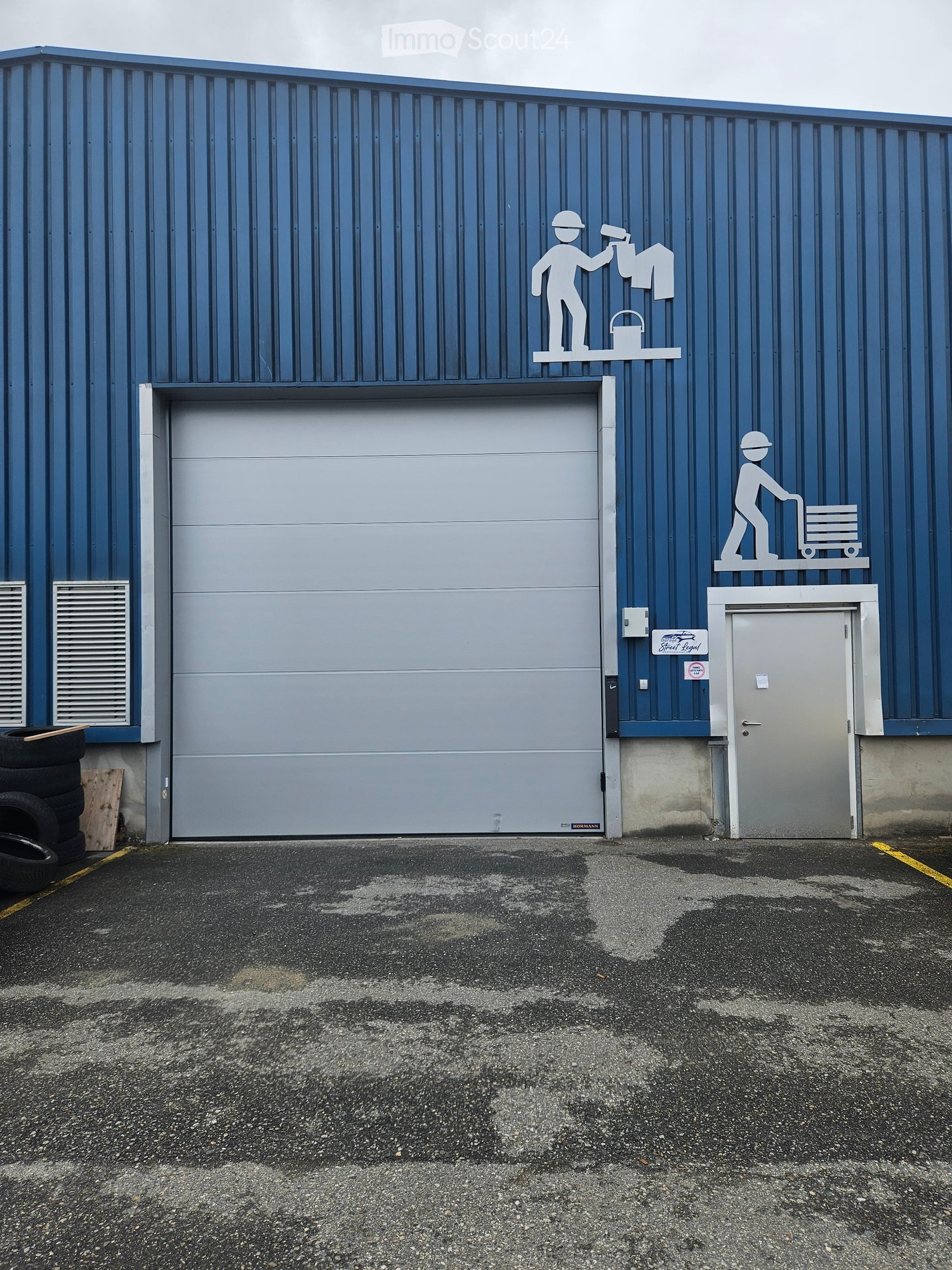 Building. A large industrial building with a blue corrugated metal exterior, featuring a large garage door, a smaller personnel entrance, and decorative wall art depicting workers.