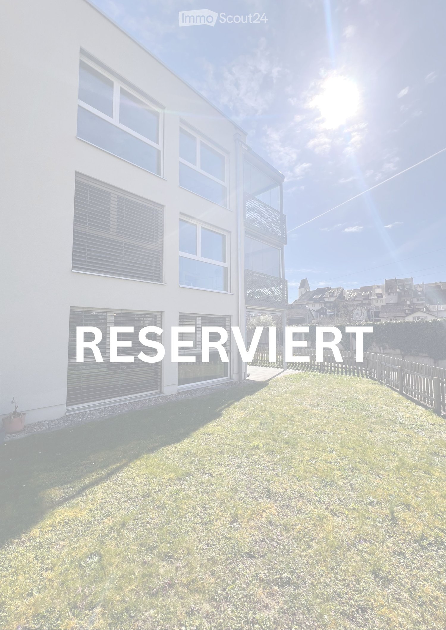 Modern white house with large windows and a balcony, surrounded by a well-maintained lawn and a small garden area.
