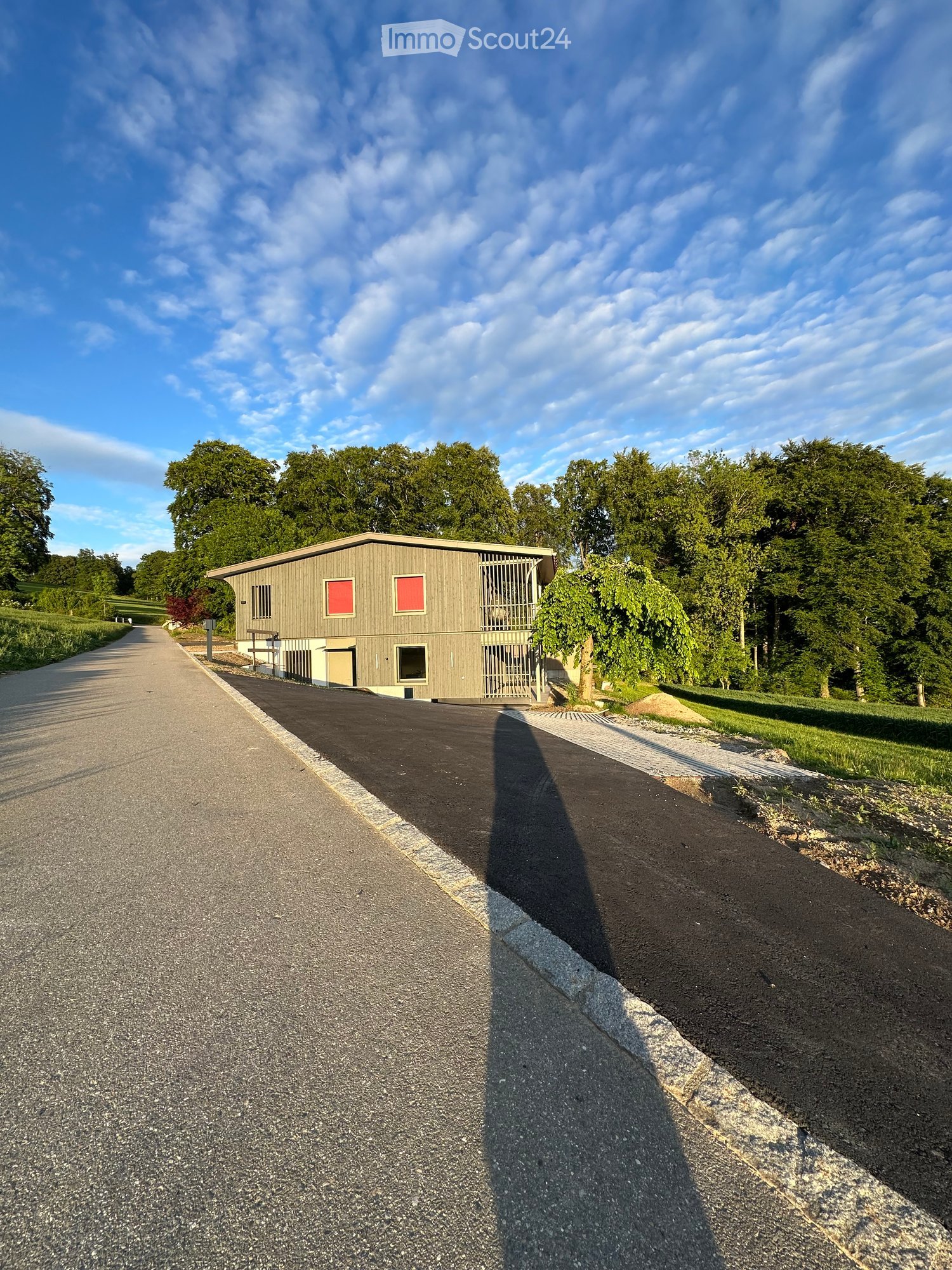 Building. A modern, single-story building with a flat roof and red-framed windows, situated on a paved driveway surrounded by greenery.