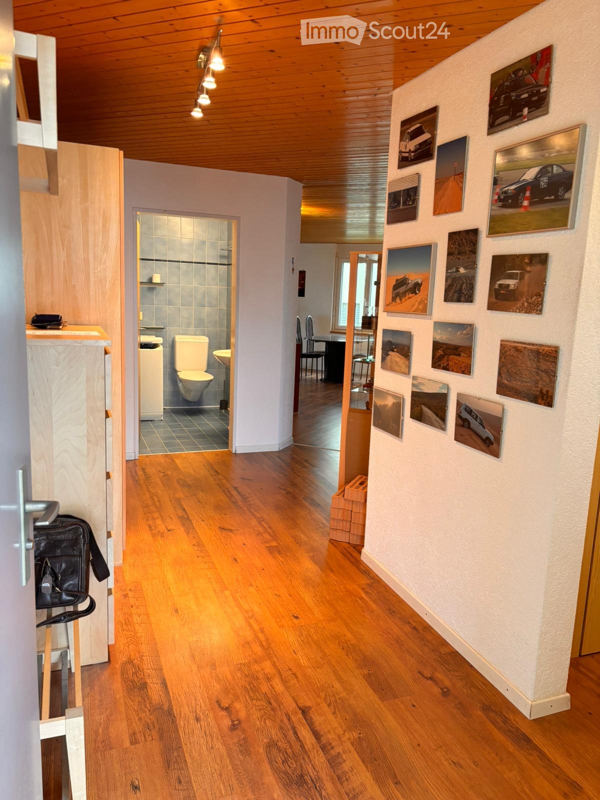 Corridor / Hallway. A well-lit hallway with wooden flooring, white walls, and several framed pictures. A small bathroom is visible to the left, and a dining area is visible to the right.