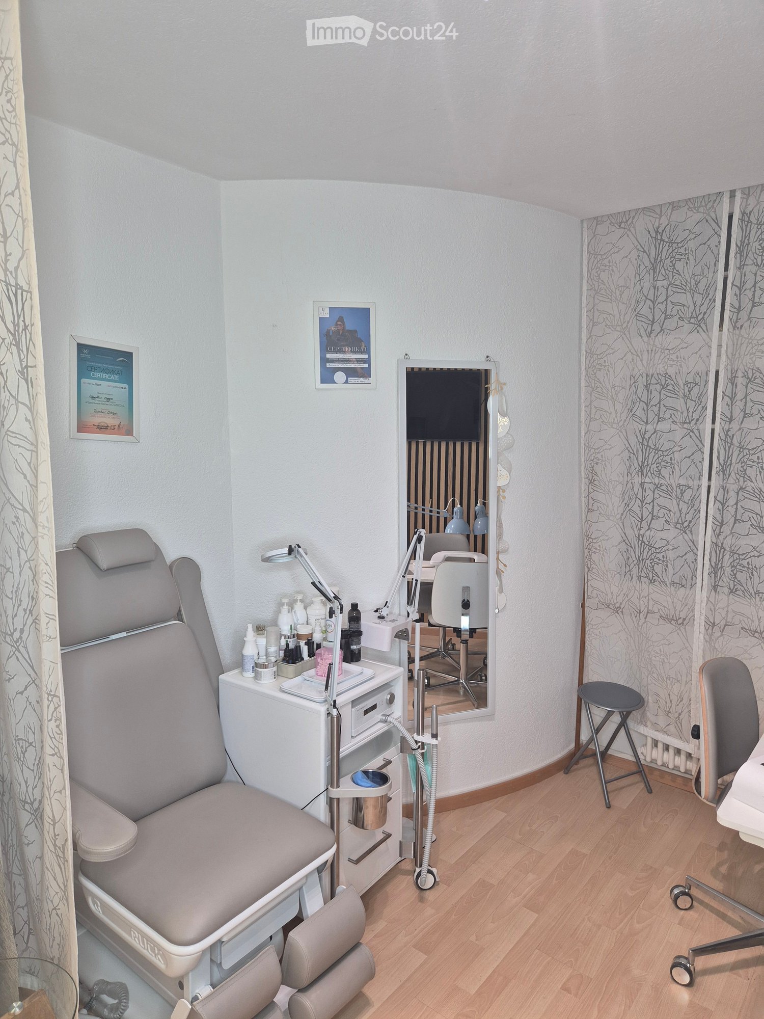Furnished Room. A well-organized beauty or treatment room with a large, comfortable treatment chair, a vanity table with various bottles and containers, a mirror, a small stool, and a lamp. The room features light-colored walls, wooden flooring, and frame