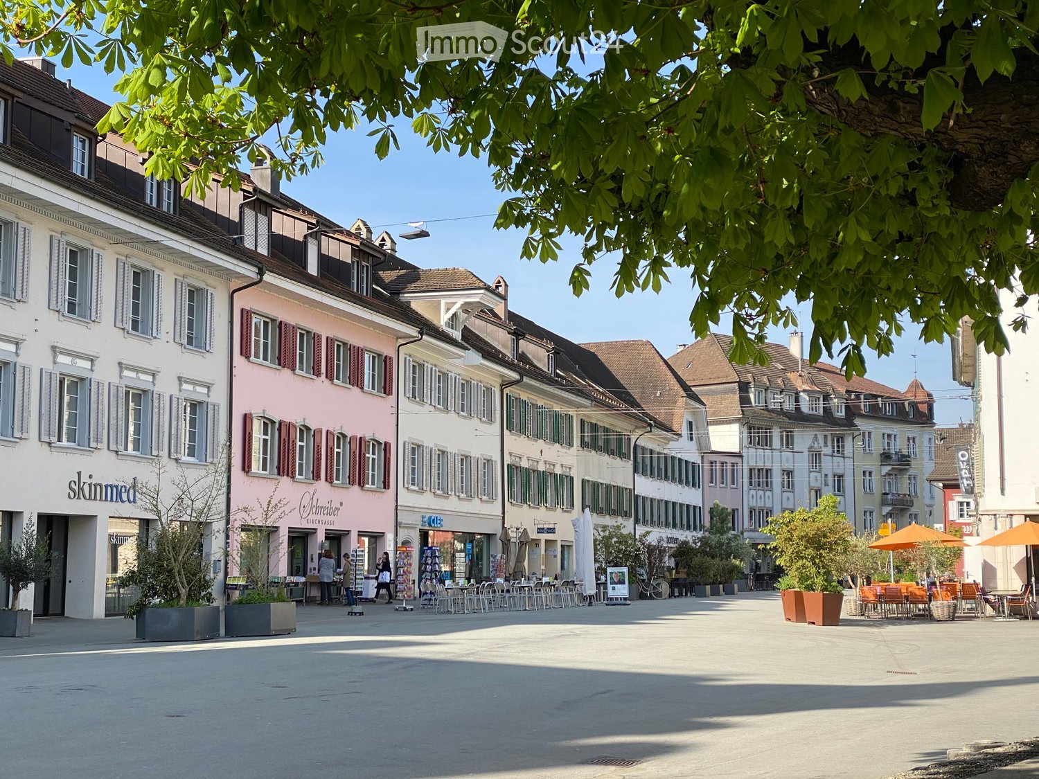 Garden / Outdoor Area. A European town square with colorful buildings, outdoor seating, and trees. The square is paved and features potted plants and umbrellas.