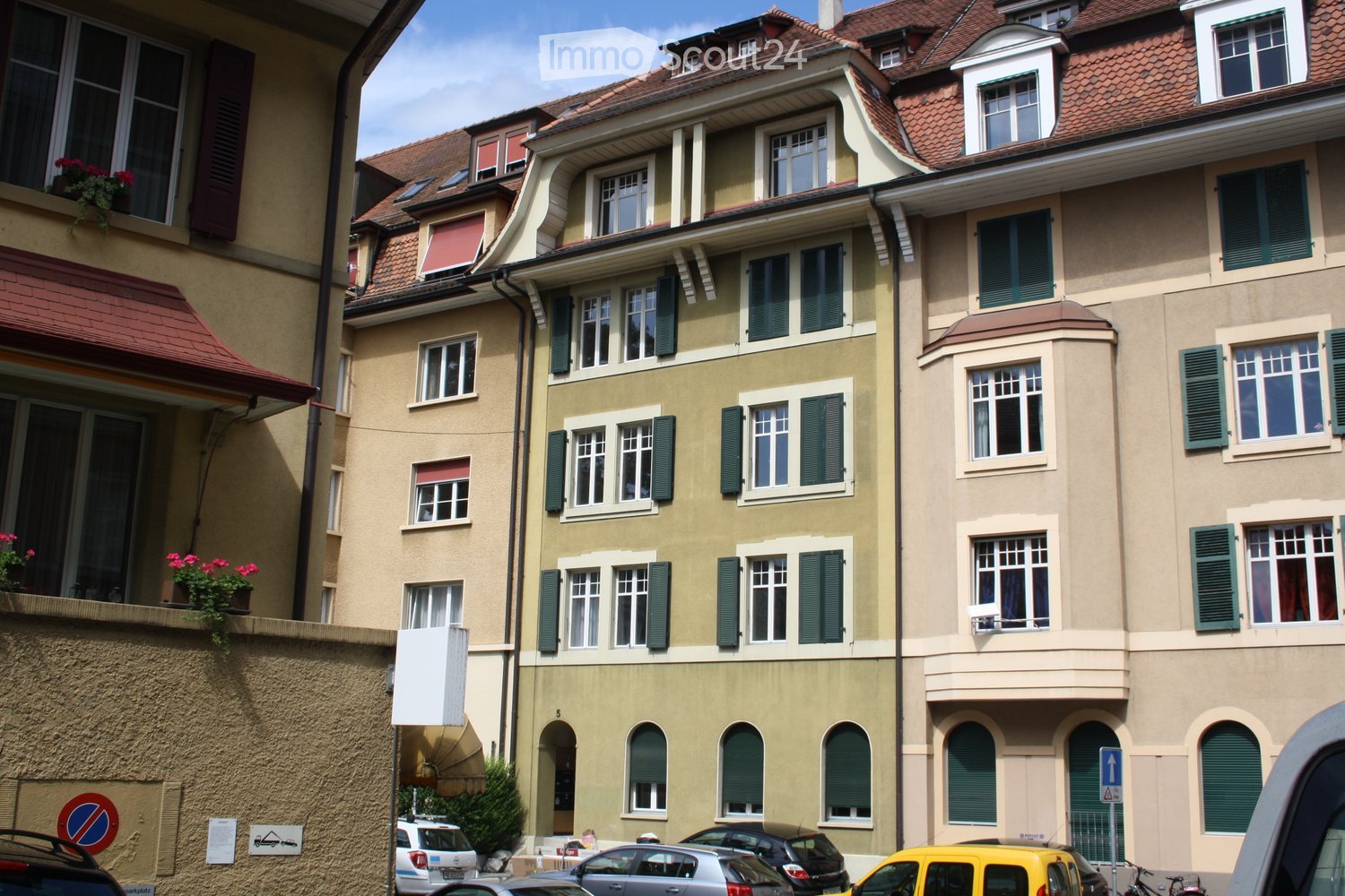Building. A multi-story residential building with beige and cream-colored facades, green shutters, and multiple windows. The building features a classic architectural style with ornate details and a red-tiled roof. There are parked cars in front of the bu