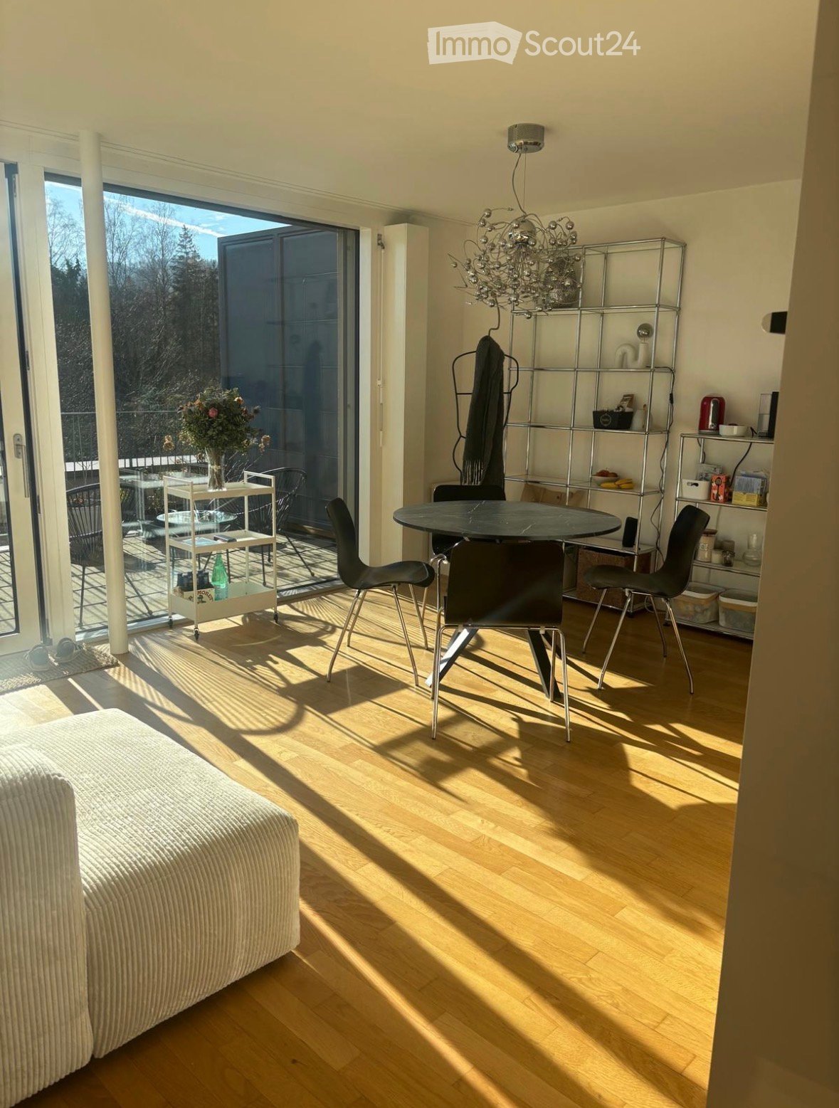 Furnished Room. Bright living and dining area with wooden flooring, large windows, a sofa, dining table with chairs, shelves, and a chandelier.