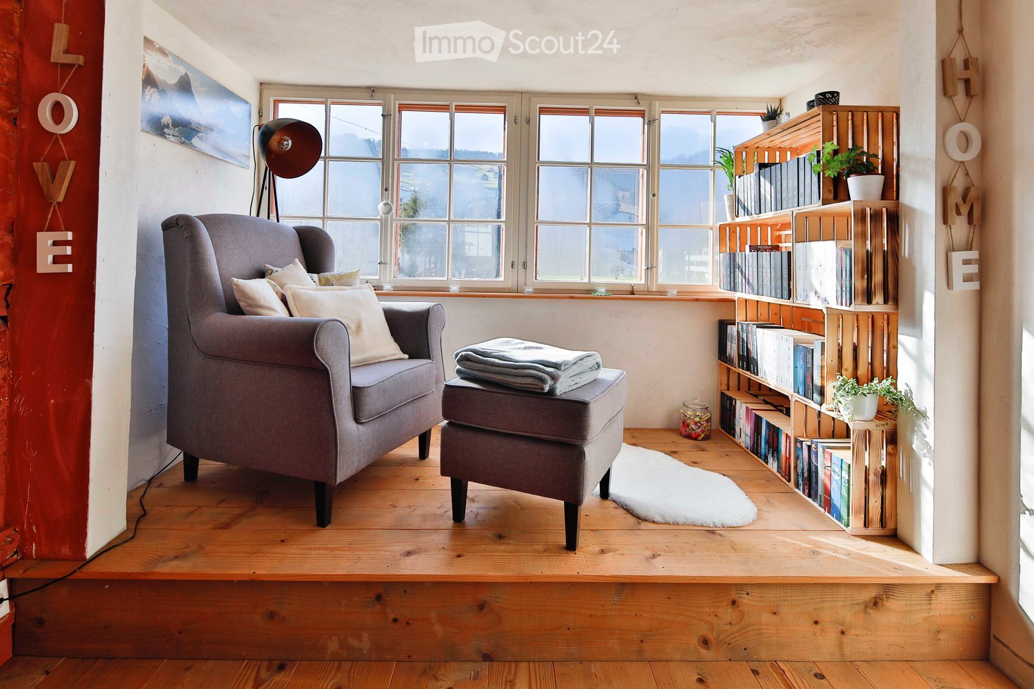 Cozy living room with wooden flooring, large windows, grey armchair, ottoman, bookshelf, floor lamp, and decorative plants.