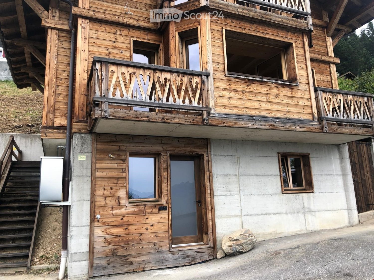 Building. A two-story wooden building with balconies, large windows, and a concrete base. The entrance features a wooden door and a staircase leading up to the upper level.