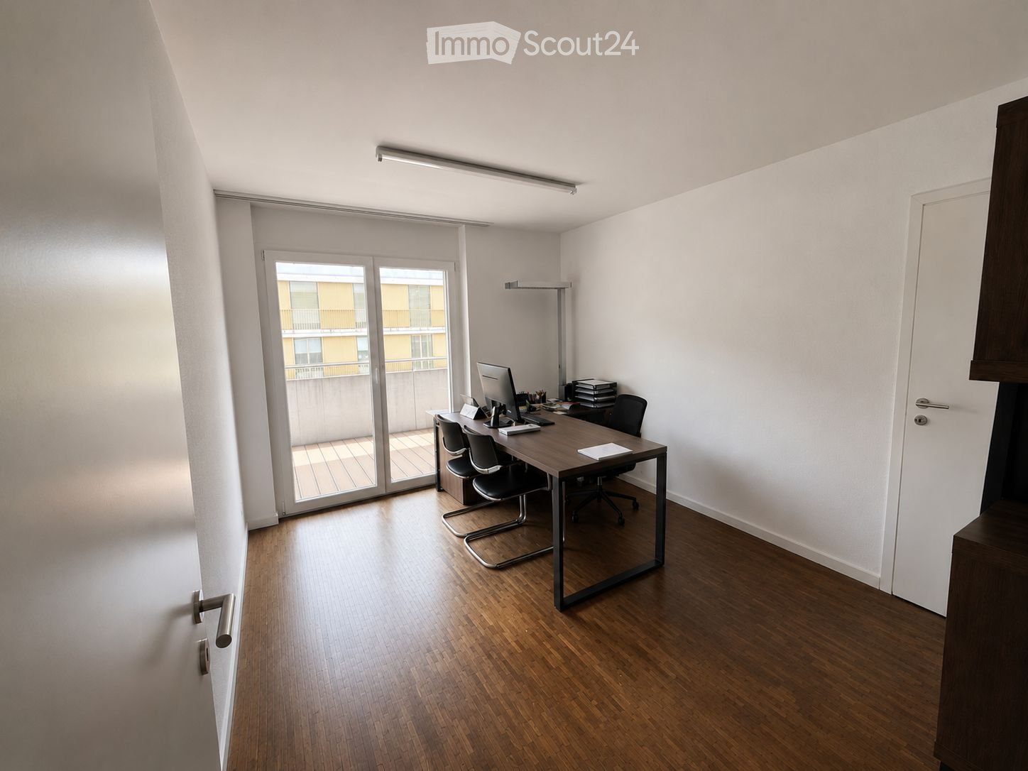 Furnished Room. A modern, well-lit office space with a wooden floor, large sliding glass doors leading to a balcony, a desk with a computer, chairs, and a lamp.