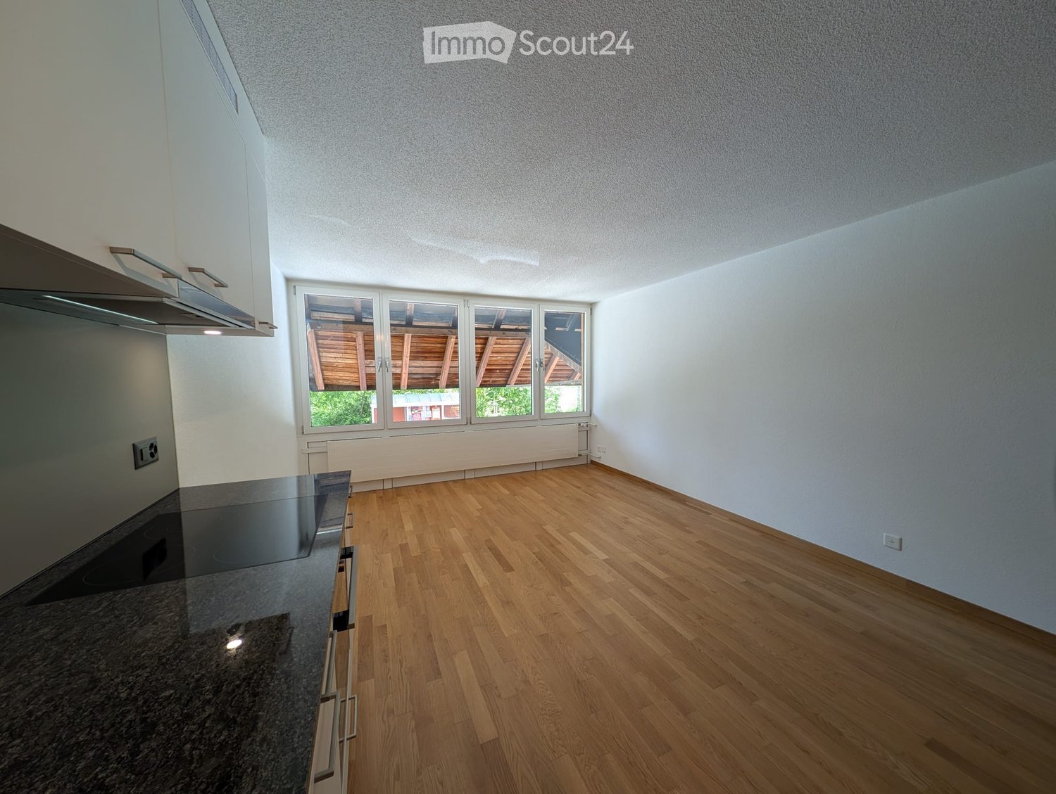 Unfurnished Room. Empty room with wooden flooring, white walls, and a large window with a view of a wooden structure outside. A modern kitchen counter is visible on the left side.