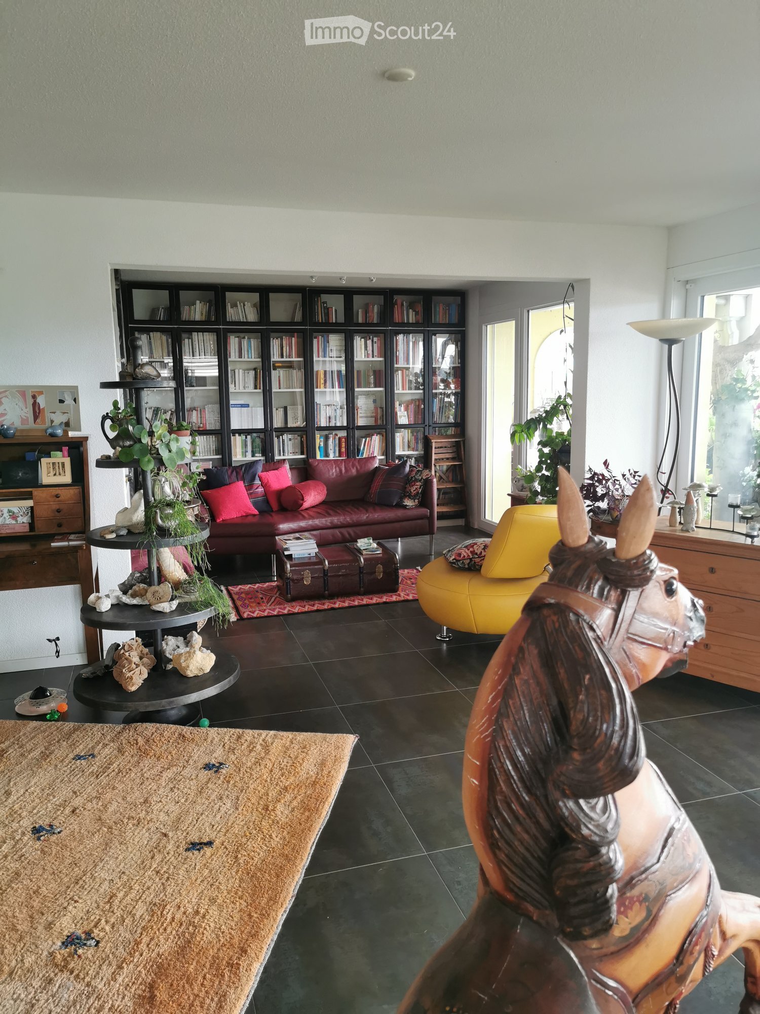 Living Room. Modern living room with dark tiled flooring, a red leather sofa, a yellow chair, a round black table, a bookshelf filled with books, and decorative items including a plant and a wooden horse statue.