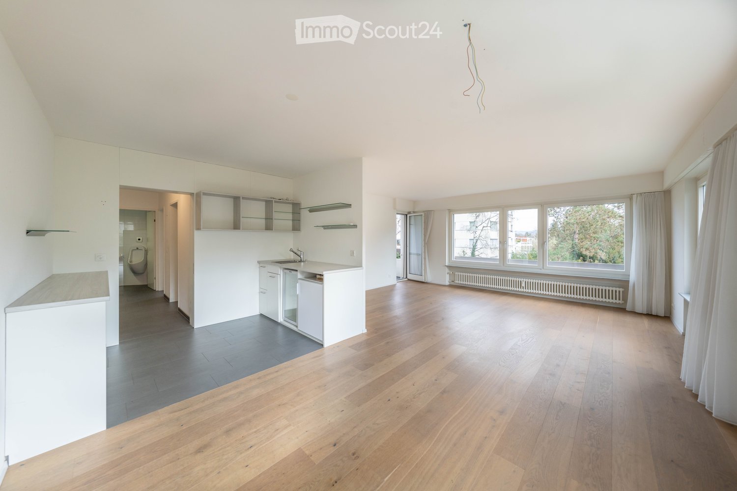 Unfurnished Room. Bright, open-plan living and dining area with wooden flooring, white walls, large windows, and a modern kitchen area with white cabinets and a sink.
