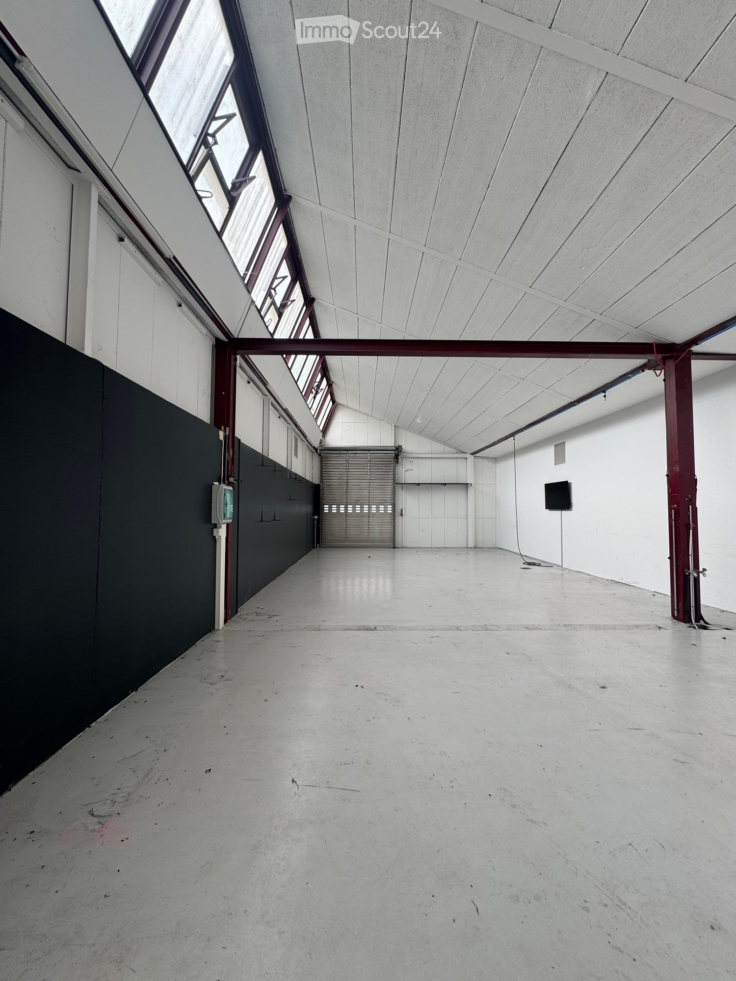 Unfurnished Room. Empty, spacious room with concrete floor, white walls, and a large garage door at the far end. The ceiling is high with exposed beams and skylights, and there is a black wall on the left side.