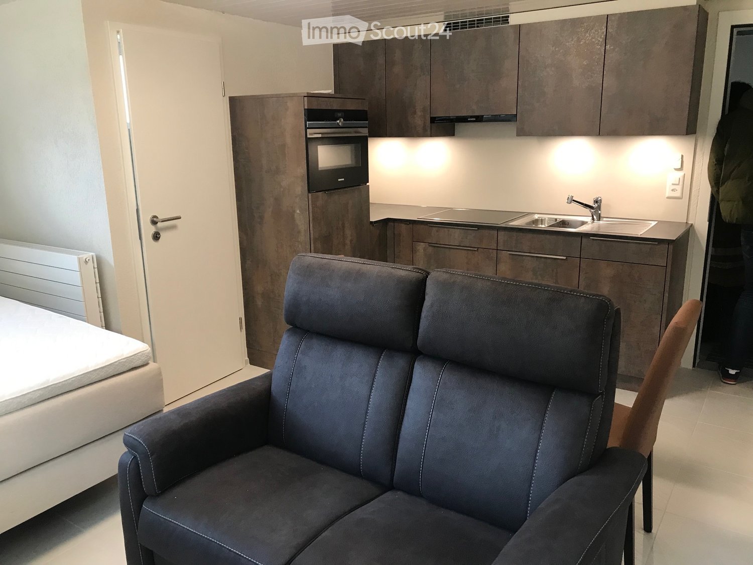 Furnished Room. Modern, compact living space with a sofa, chair, bed, and a small kitchen area featuring cabinets, a sink, and an oven.